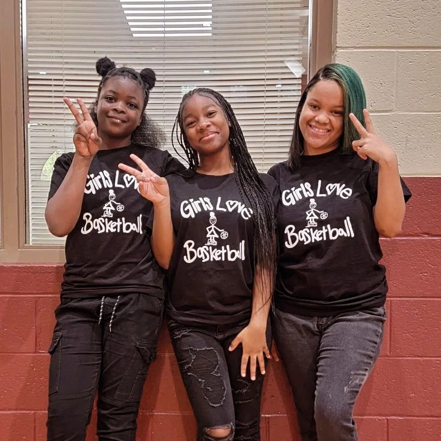 Three young girls standing against a brick wall, each wearing a black T-shirt that says "Girls Love Basketball" and making peace sign gestures.