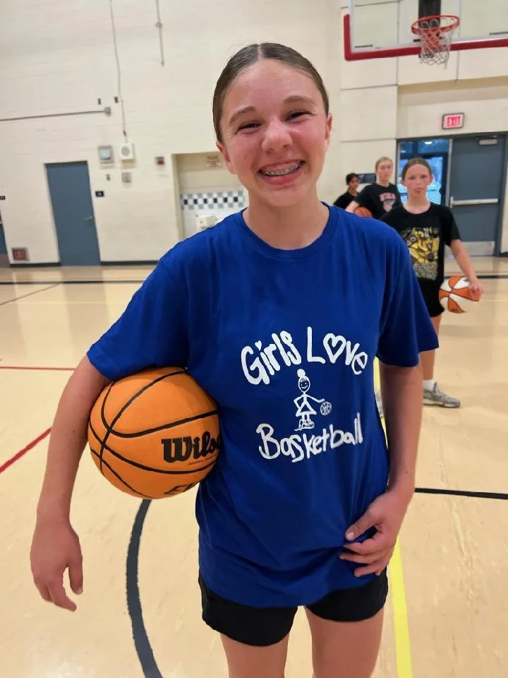 A girl smiling and holding a basketball in a gymnasium, wearing a blue t-shirt that says 'Girl Love Basketball'.