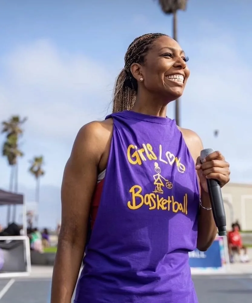 Woman smiling and holding a microphone outdoors, wearing a purple shirt that says "Girl Love Basketball," with palm trees and a clear sky in the background.