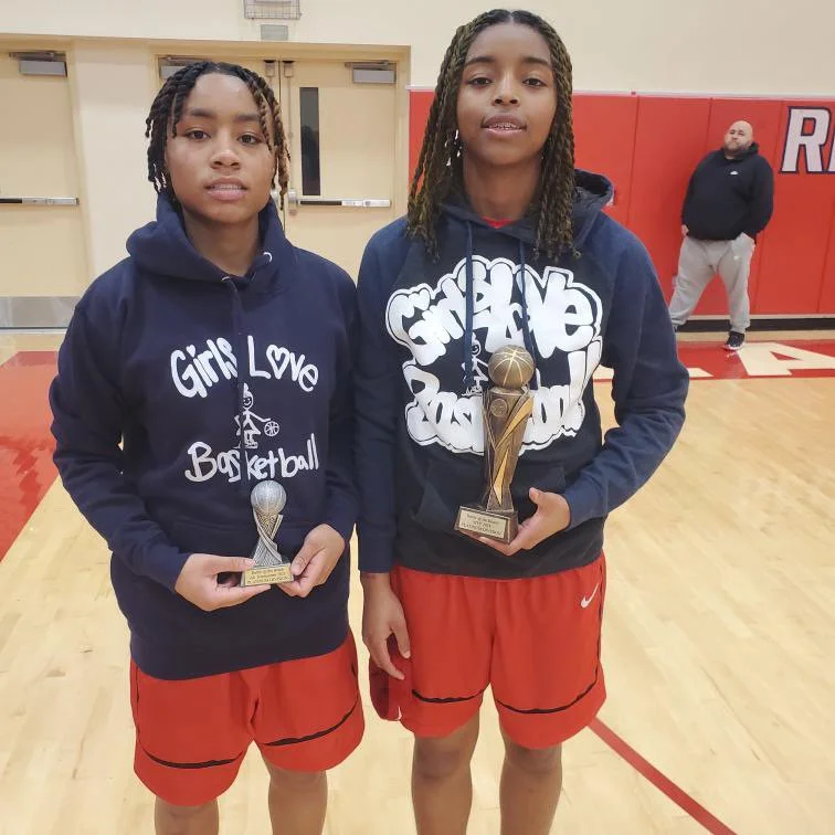 Two young girls wearing navy blue hoodies and red athletic shorts standing on a basketball court, each holding a trophy. The hoodies say 'Girls Love Basketball'.