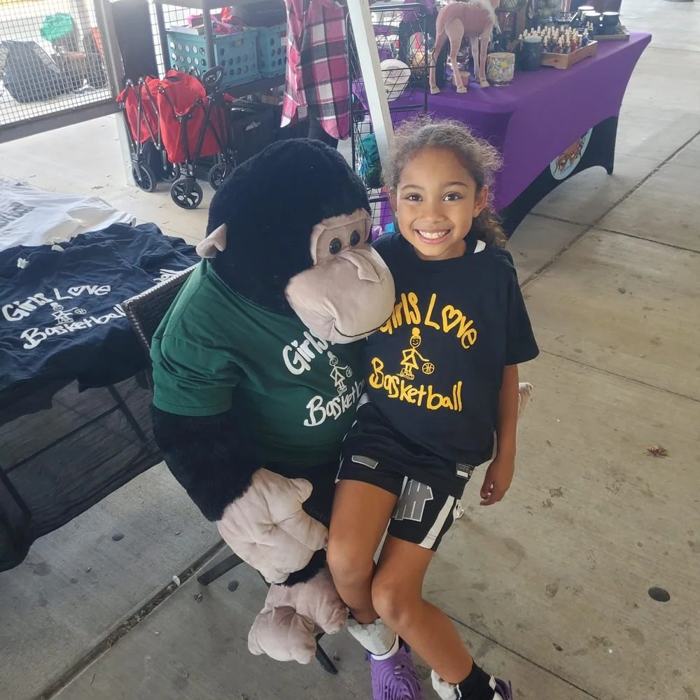 A young girl sitting with a big stuffed gorilla, both smiling at the camera. They are at a booth with various items displayed, including t-shirts with 'Girls Love Basketball' printed on them.