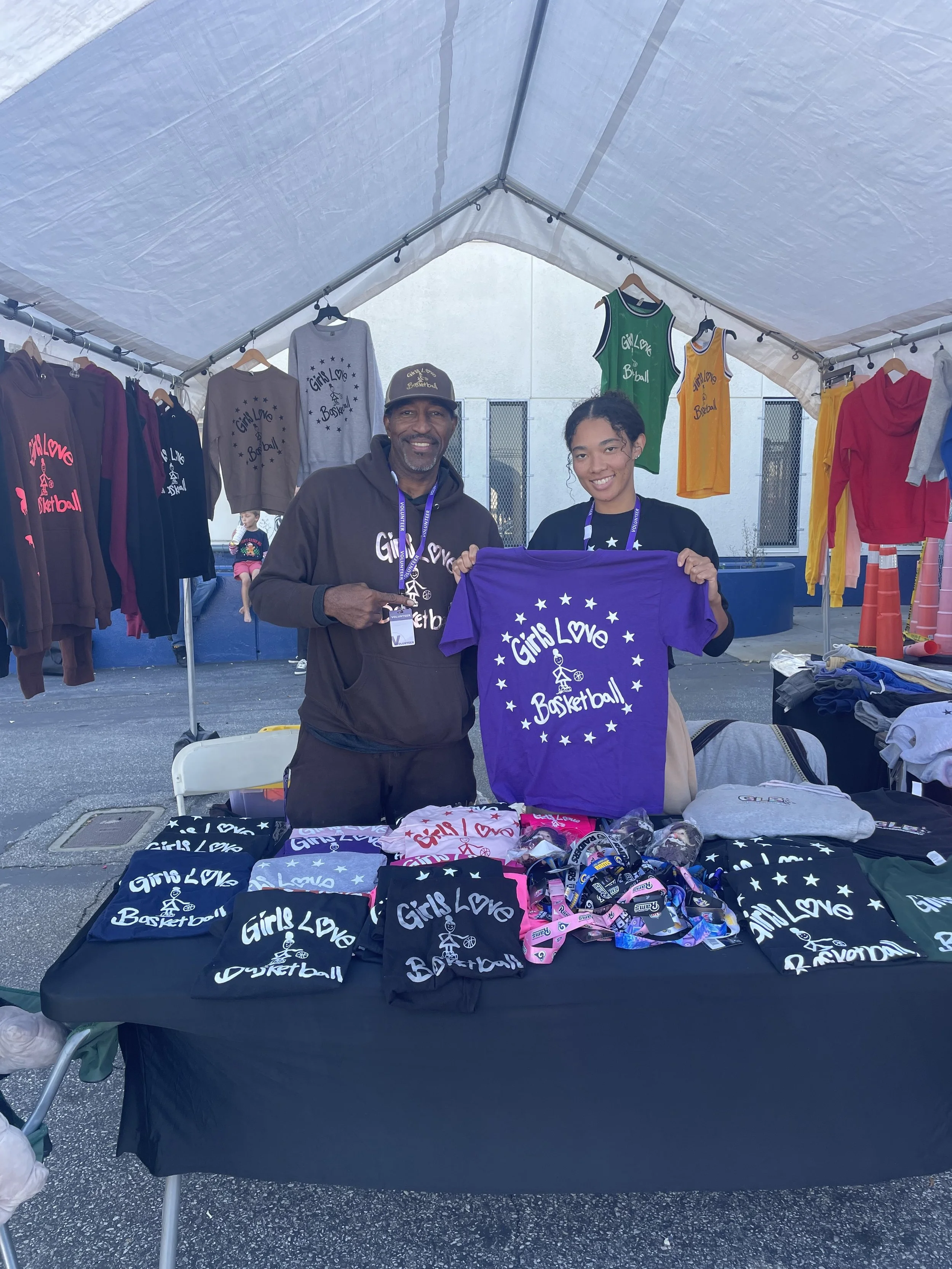 A woman and a man are standing behind a table at a booth selling merchandise. The table has shirts and wristbands with the slogan 'Girls Love Basketball.' They are holding a purple shirt with the same slogan. The booth displays various hoodies and ta