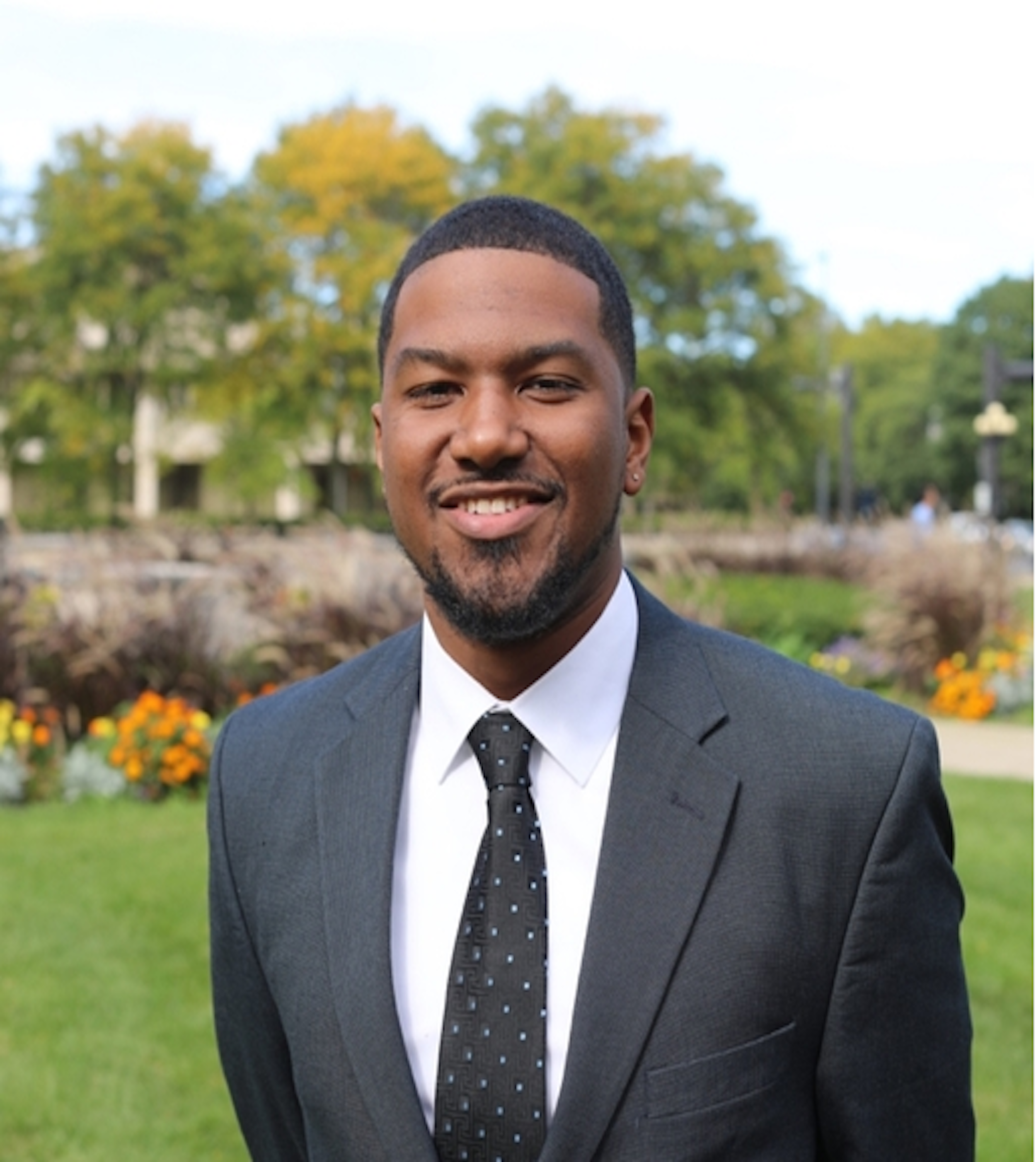 Portrait of a smiling man in a suit outdoors with trees and flowers in the background.