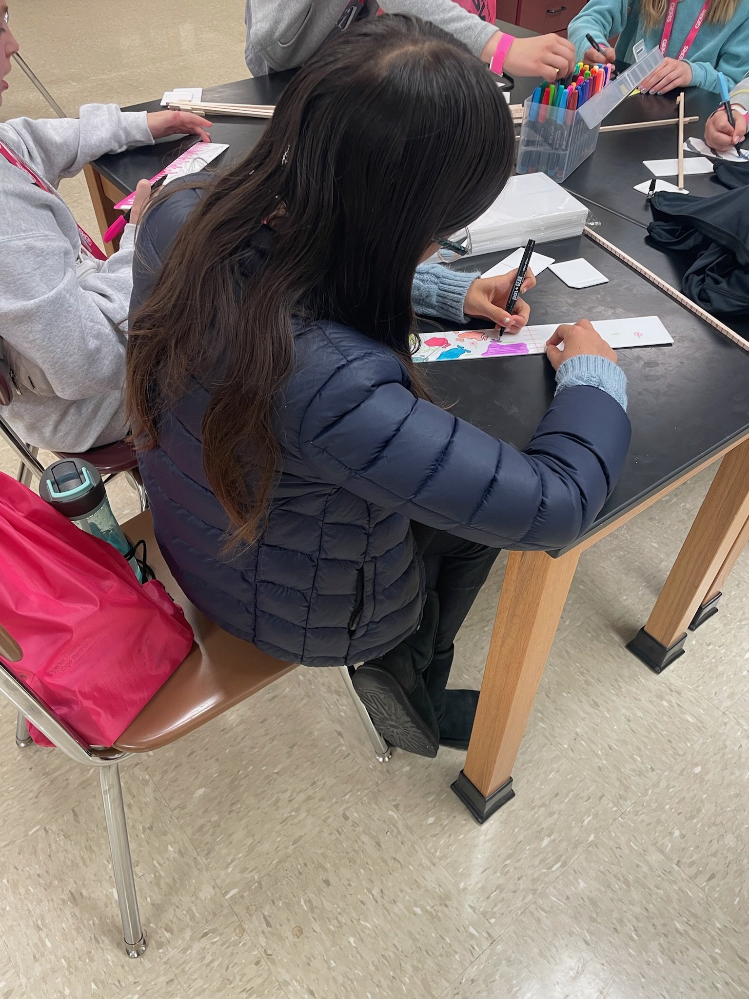 A girl with long brown hair, wearing glasses, a blue jacket, and black pants, sitting at a table drawing on a strip of paper with markers. Other children are also seated at the table, engaging in similar activities, with colorful markers and supplies on the table.