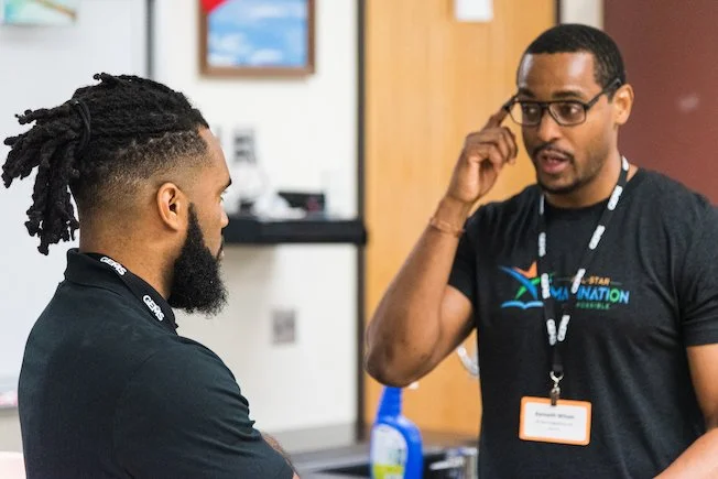 Two men having a conversation indoors, one is wearing glasses and a black shirt with a lanyard, the other has dreadlocks and a beard, also wearing a black shirt with a lanyard.