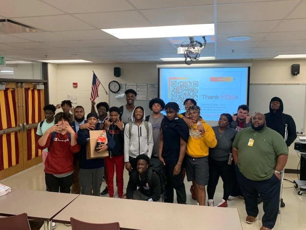 Group of students and teachers posing for a photo in a classroom, with a QR code and a Thank You message on the screen in the background.