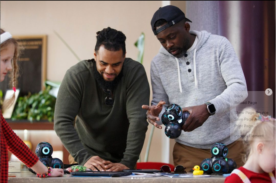 Two men and a young girl play with small black robots with glowing blue accents on a table in a room.