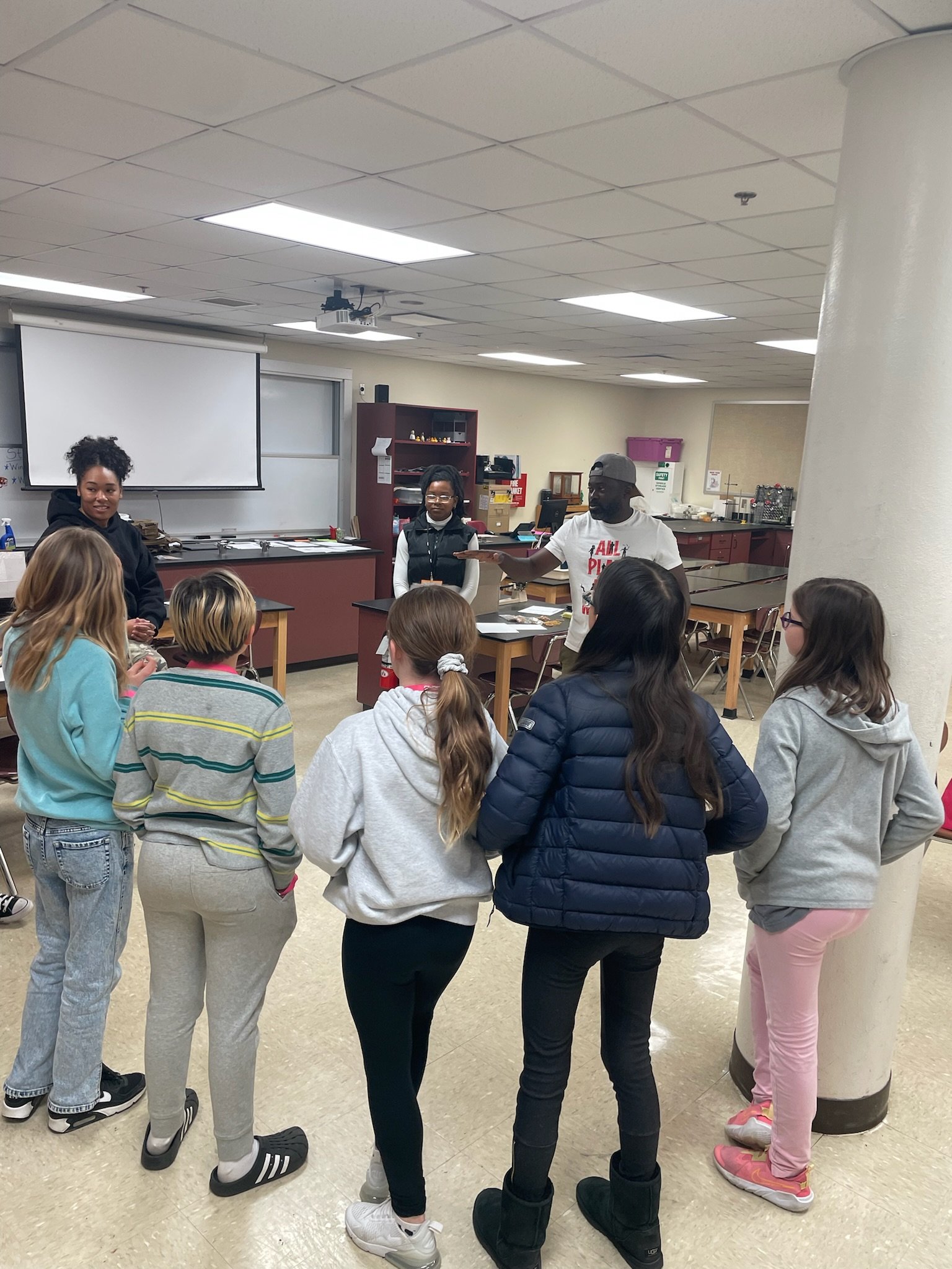 A group of young students standing in a classroom, listening to a man speaking, with a woman nearby. The classroom has a whiteboard, shelves, and tables.