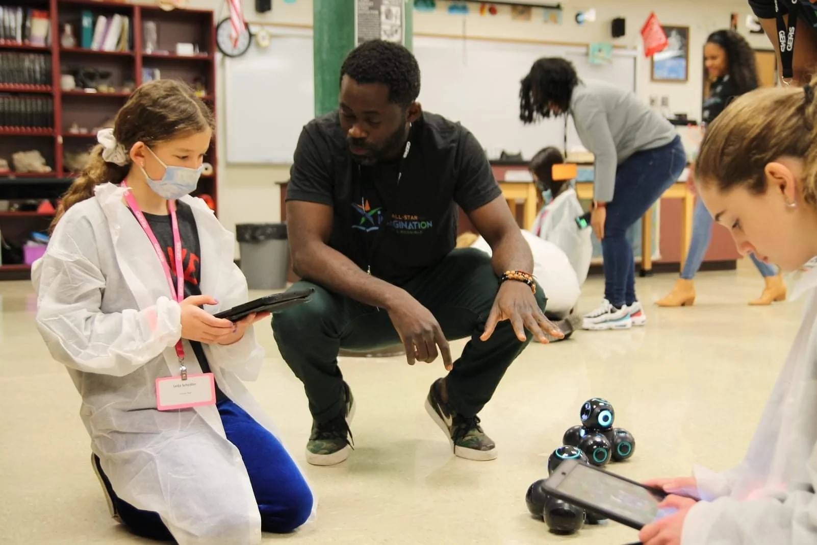 Children and an adult interacting with small robots in a classroom. A girl in a white coat and face mask looks at a tablet, while another girl in a similar coat holds a tablet. The man in the middle observes, with other children and educators engaged in activities in the background.