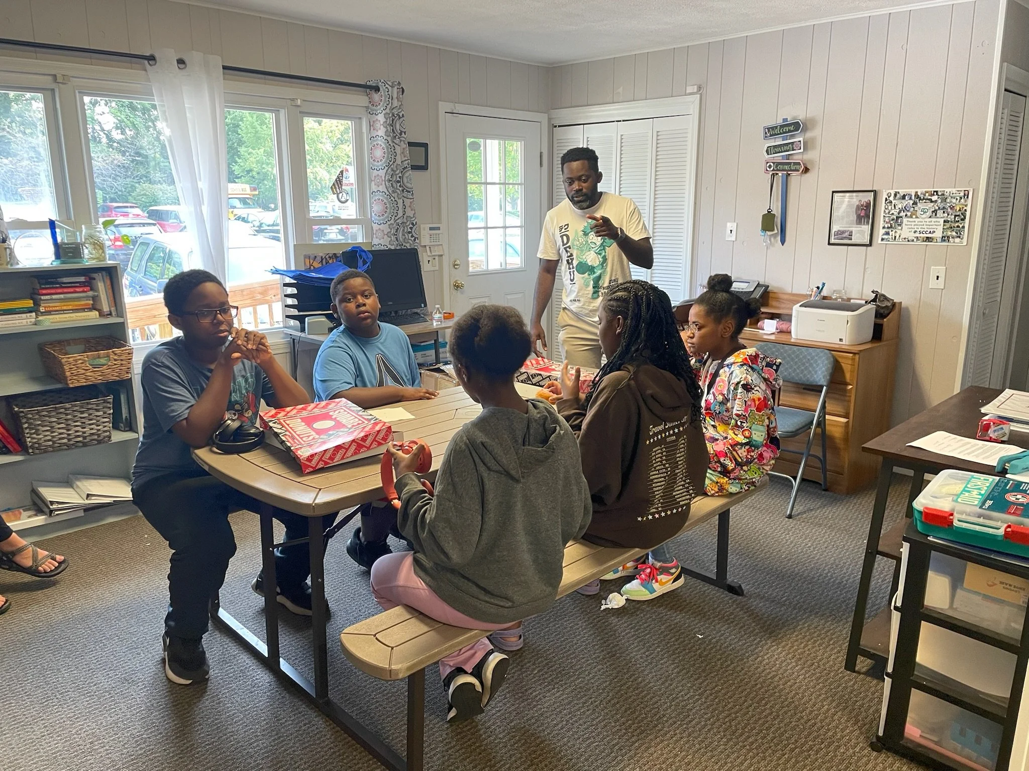 A group of children sitting at a table in a classroom, listening to an instructor who is standing and gesturing. The classroom has windows, shelves with books, and a bulletin board on the wall.