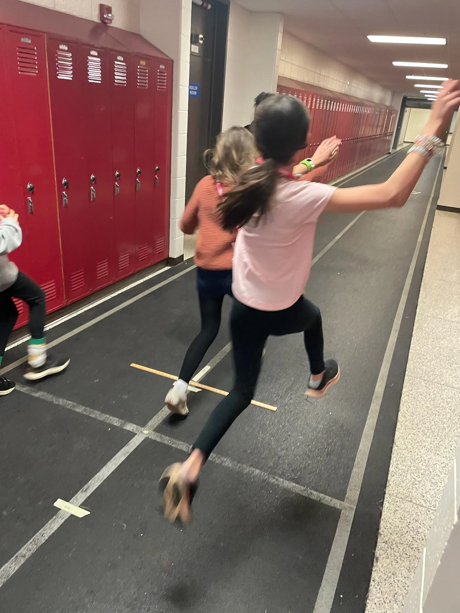 Two students running in a school hallway, with red lockers along the wall and a black floor.