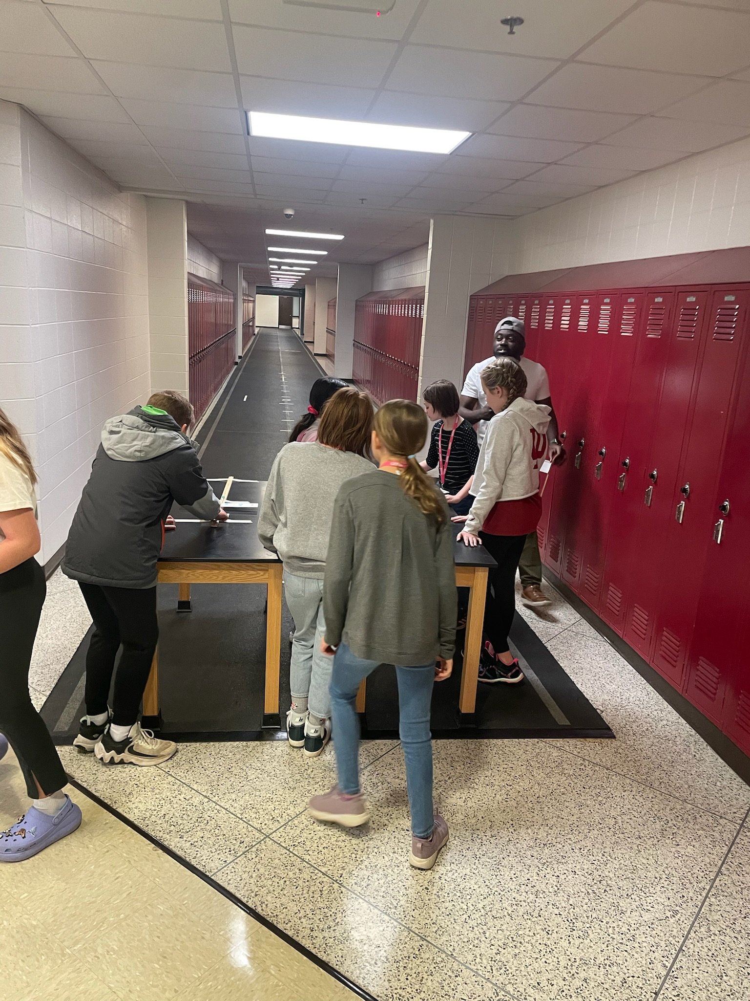 A group of students gathered around a lab table in a school hallway, with some writing and others observing, and a teacher or instructor demonstrating something.