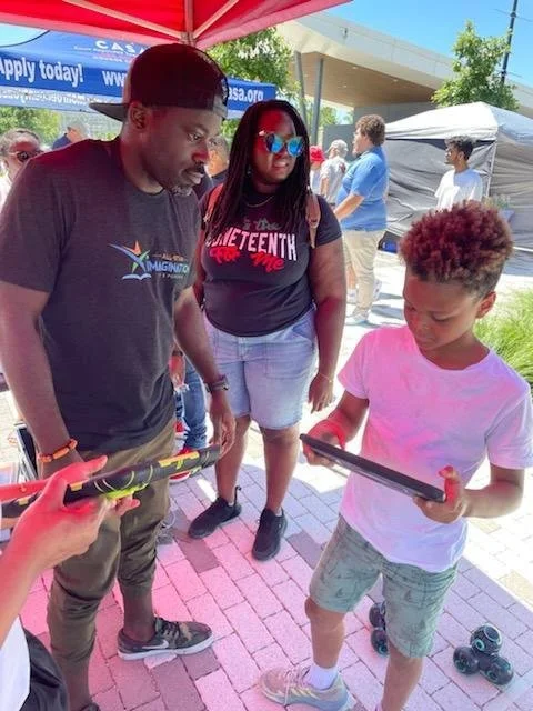 A young boy with curly hair wearing a pink t-shirt and shorts, holding a tablet, is looking at it. Two adults, a man in a black t-shirt and a woman in a black t-shirt with red and white text, are standing nearby, observing the boy. The scene is outdoors at an event with tents and other people in the background.