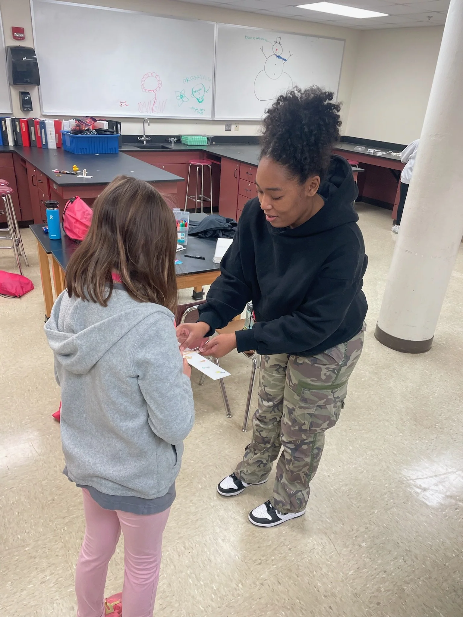 A woman in a black hoodie handing a card or paper to a young girl in a grey hoodie and pink pants in a classroom setting.