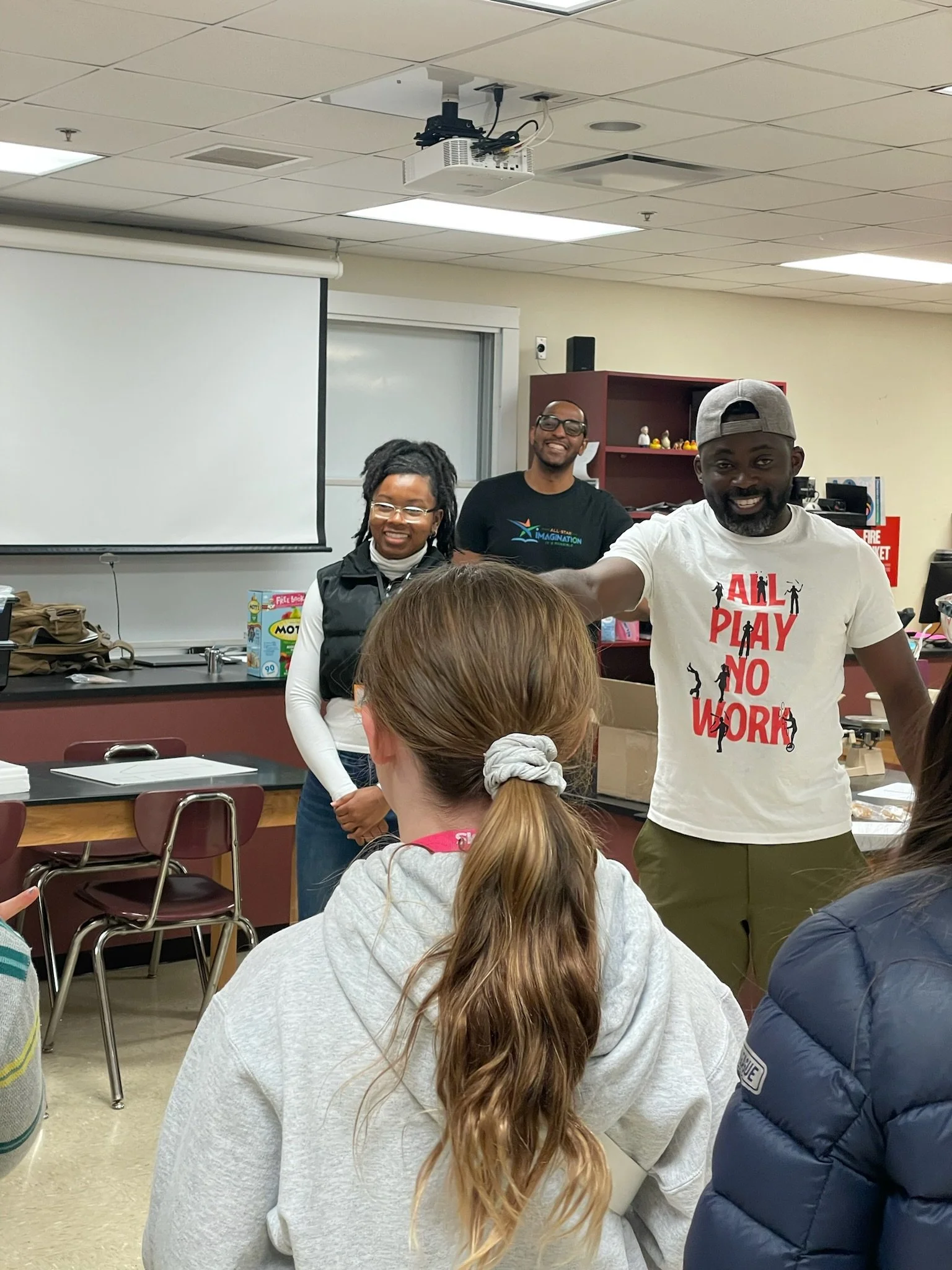 Three people standing at the front of a classroom, smiling, while students sit and face them. The classroom has a white projection screen and a reddish-brown shelf with various items.