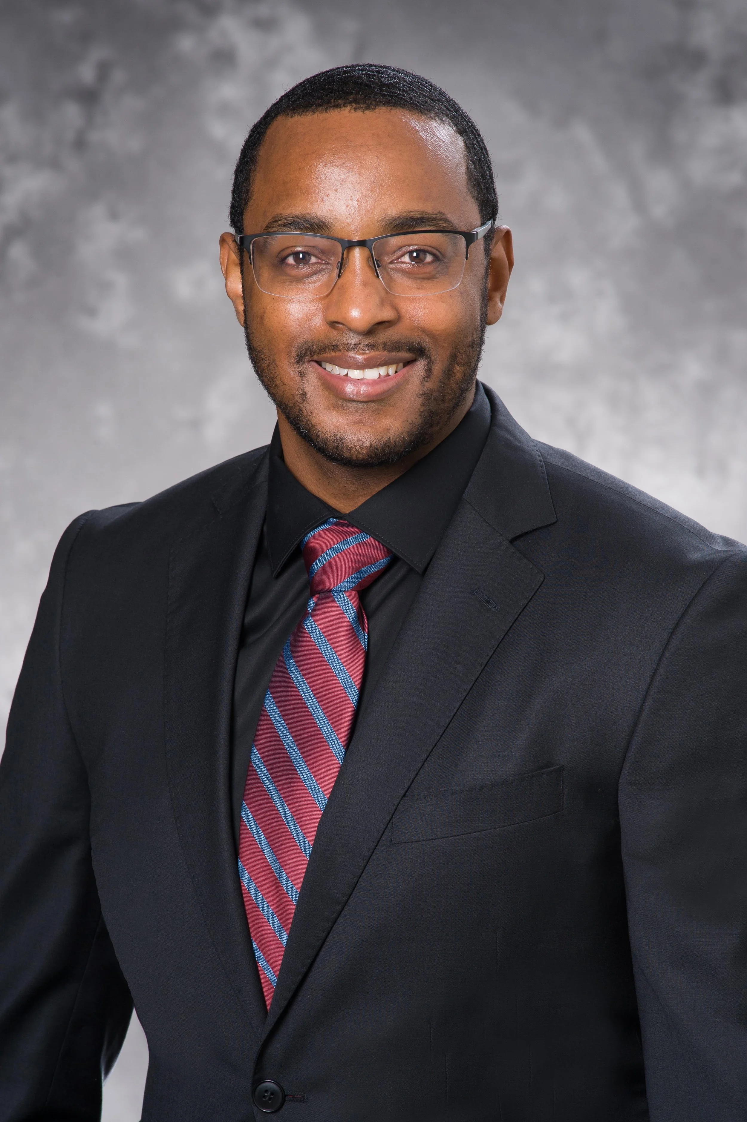 A man in a black suit, striped maroon and blue tie, and glasses, smiling in front of a gray background.
