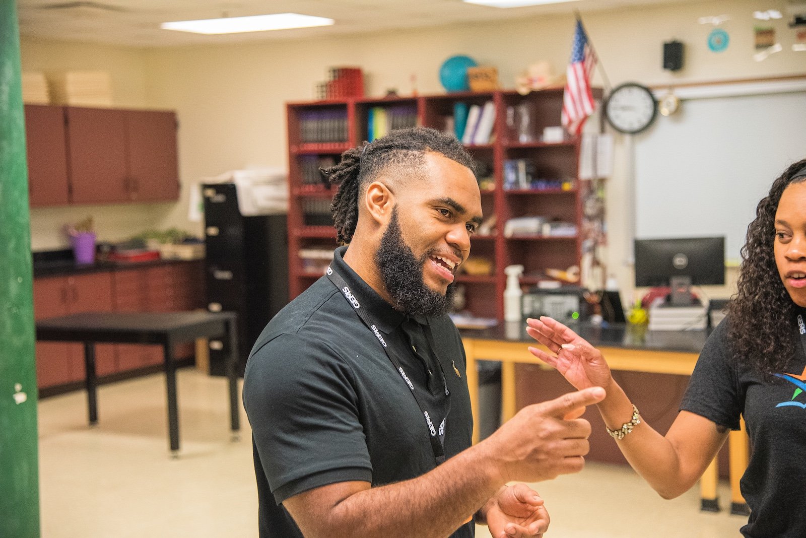 A man with a beard and dreadlocks smiling and gesturing with his hand while talking to a woman in a classroom or office environment.