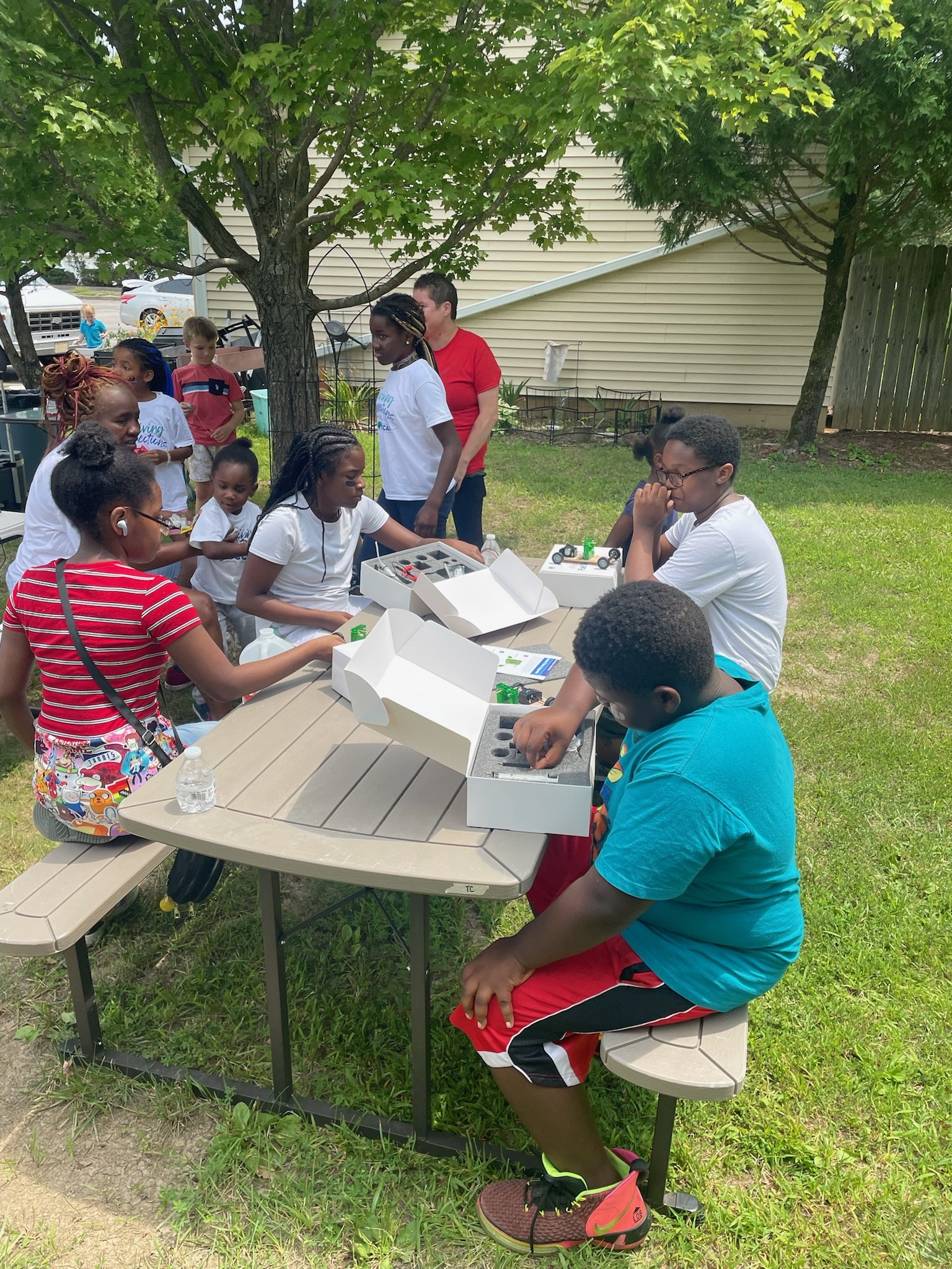 A group of children and adults gathered around a table outdoors, with some of the children working on science or engineering projects with kits, on a grassy area under trees.
