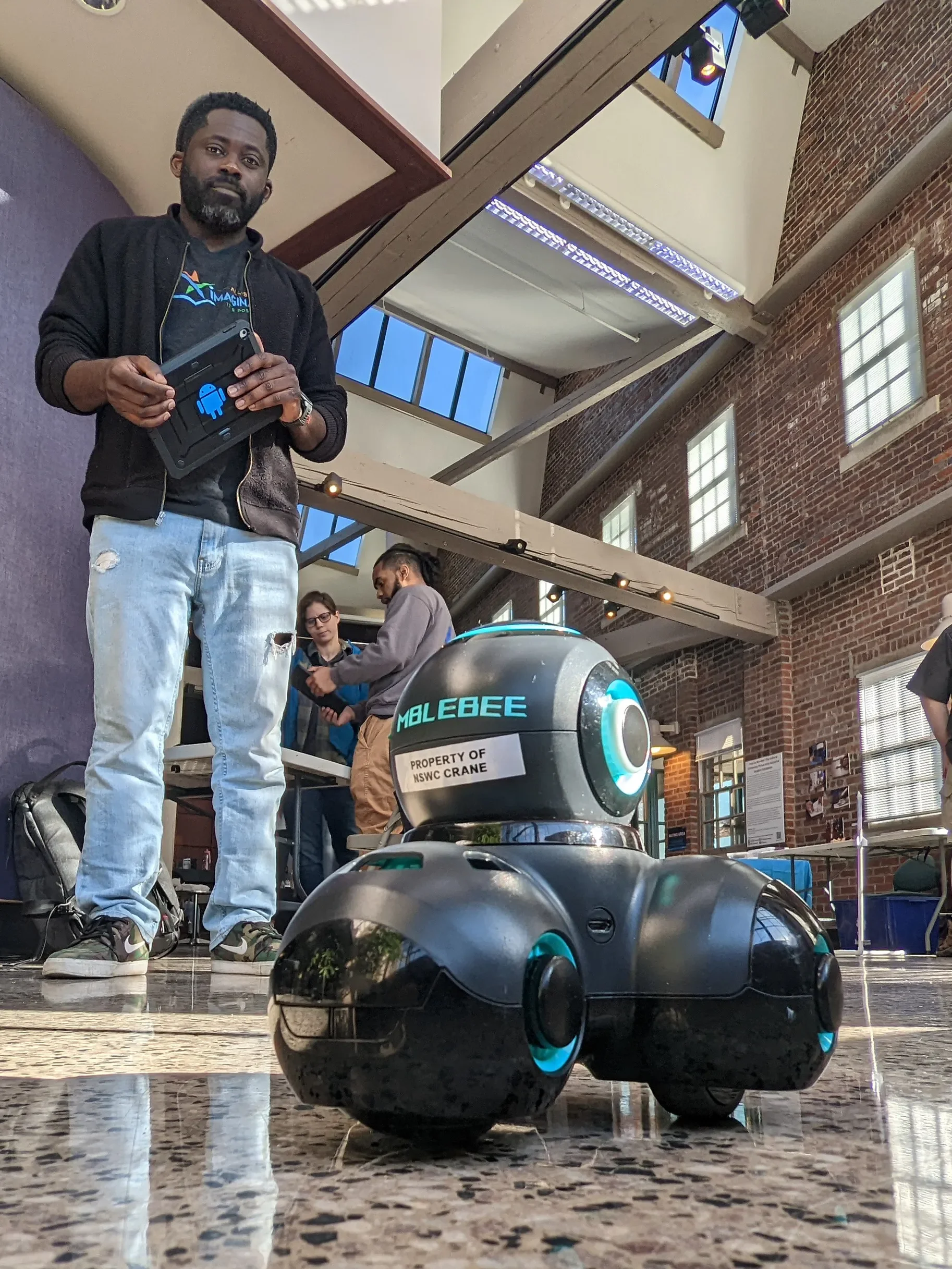 A man standing in an indoor setting holding a tablet with the Android logo on it. In front of him is a small, black, four-wheeled robot with blue accents. The background shows other people and a brick wall with windows.