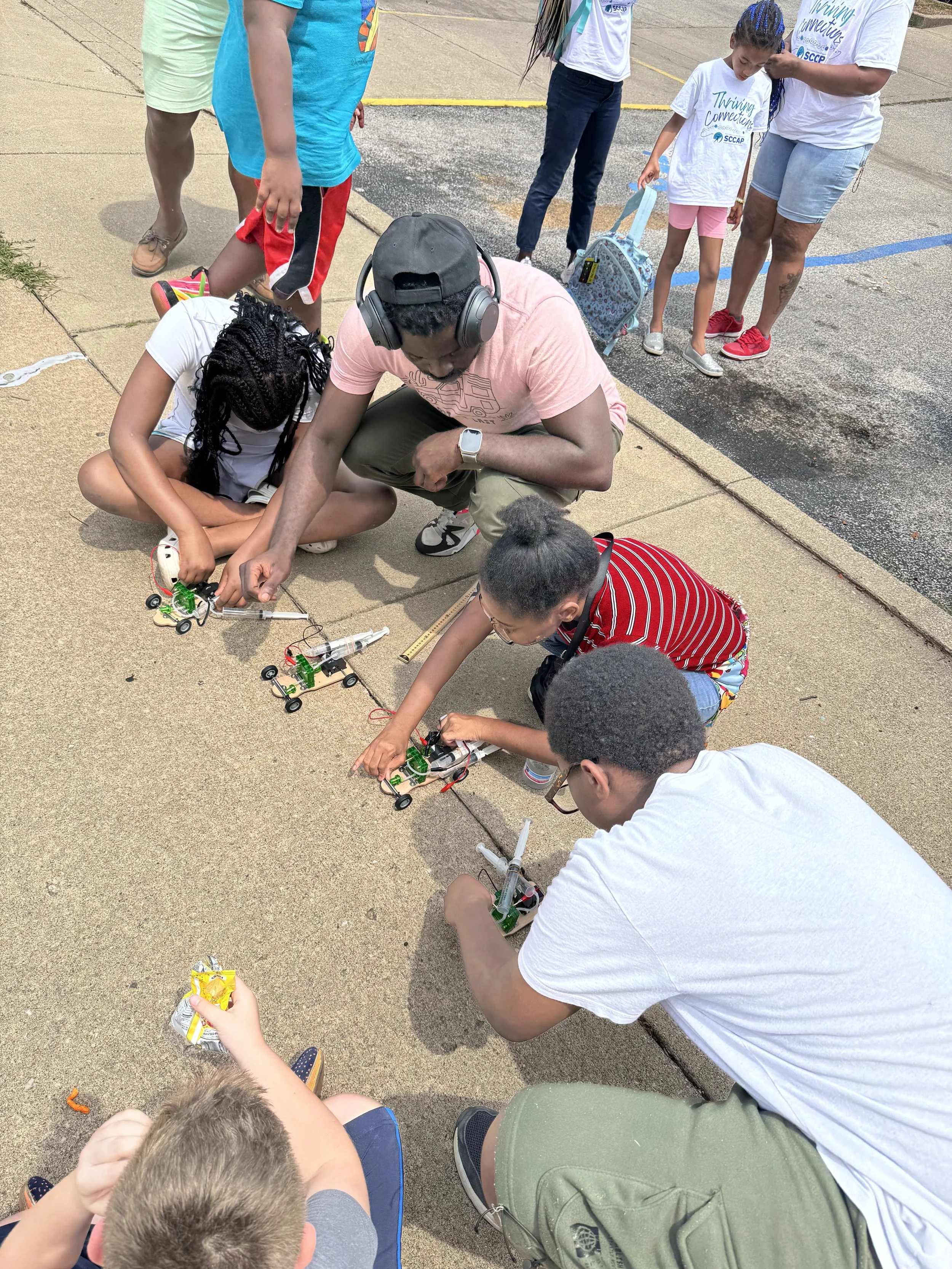 Group of children and adults outside on pavement, working on small robotic vehicles, with some standing and others crouched down, demonstrating a hands-on STEM activity.