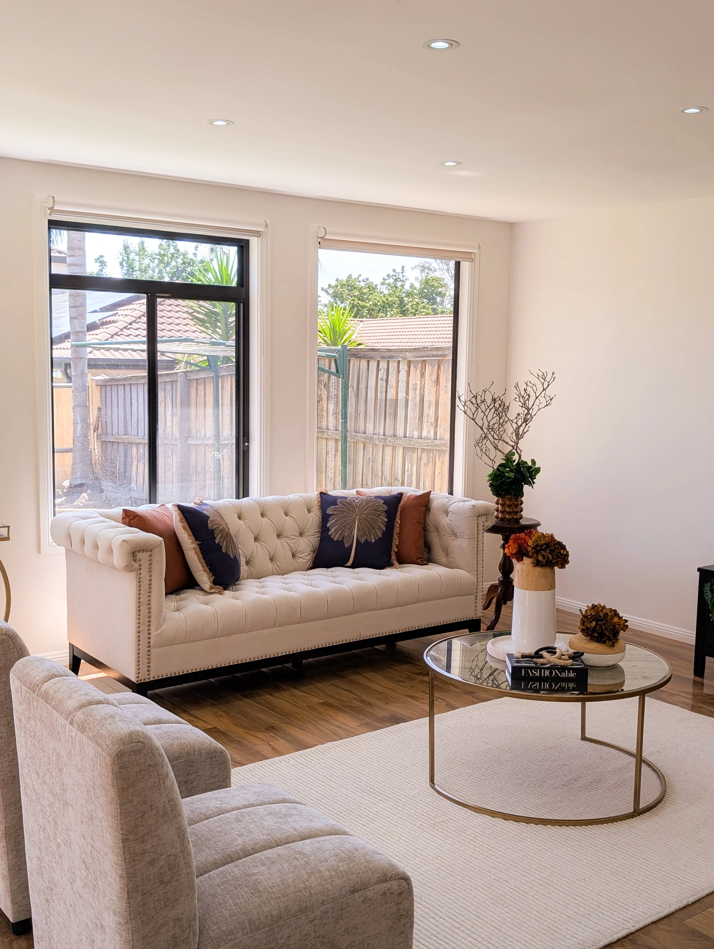 Living room with white tufted sofa, beige armchair, round glass coffee table, and large windows with outside view of a wooden fence and trees.