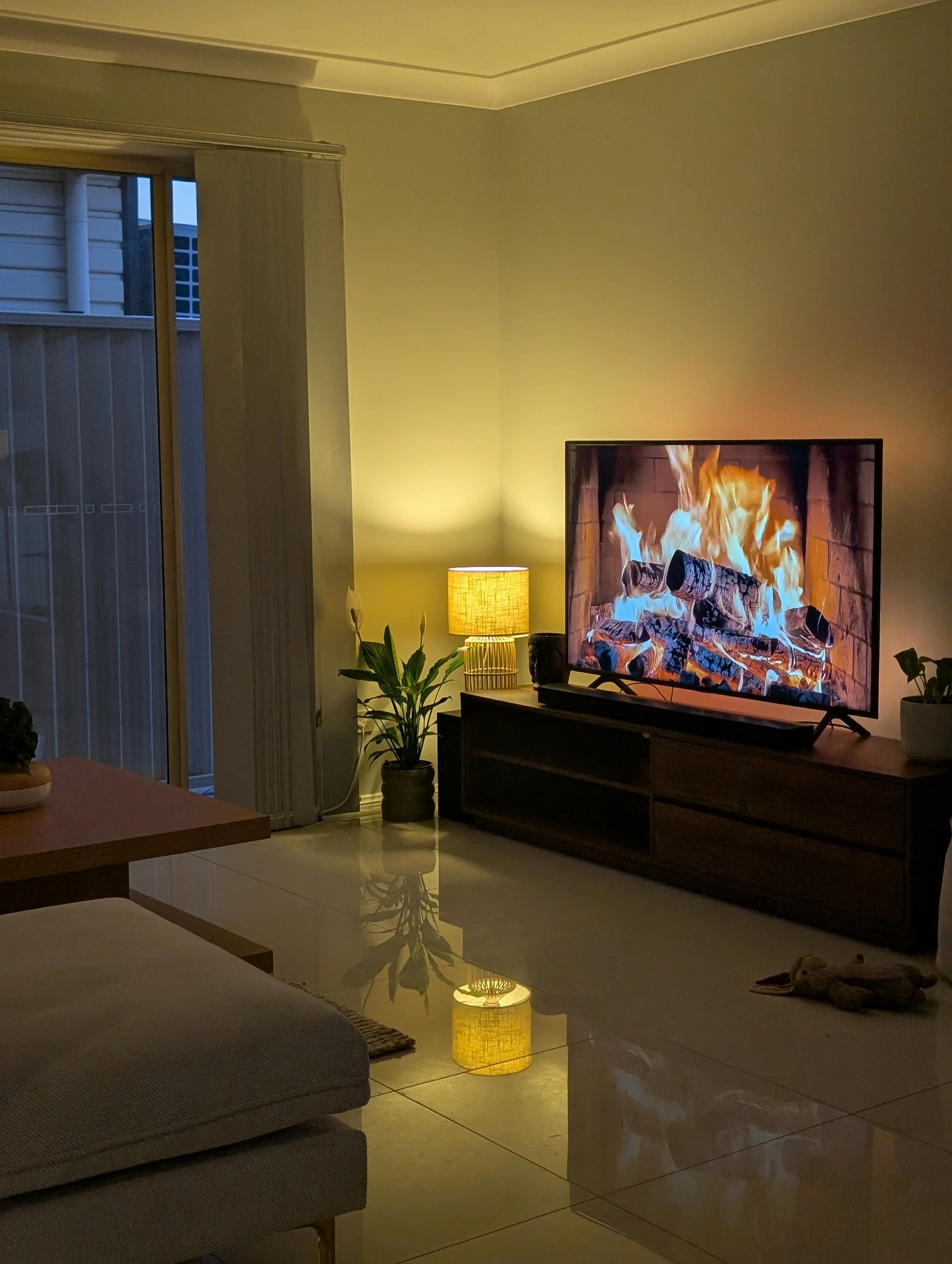 Living room with a TV displaying a fireplace, a floor lamp, a potted plant, and sliding glass doors.