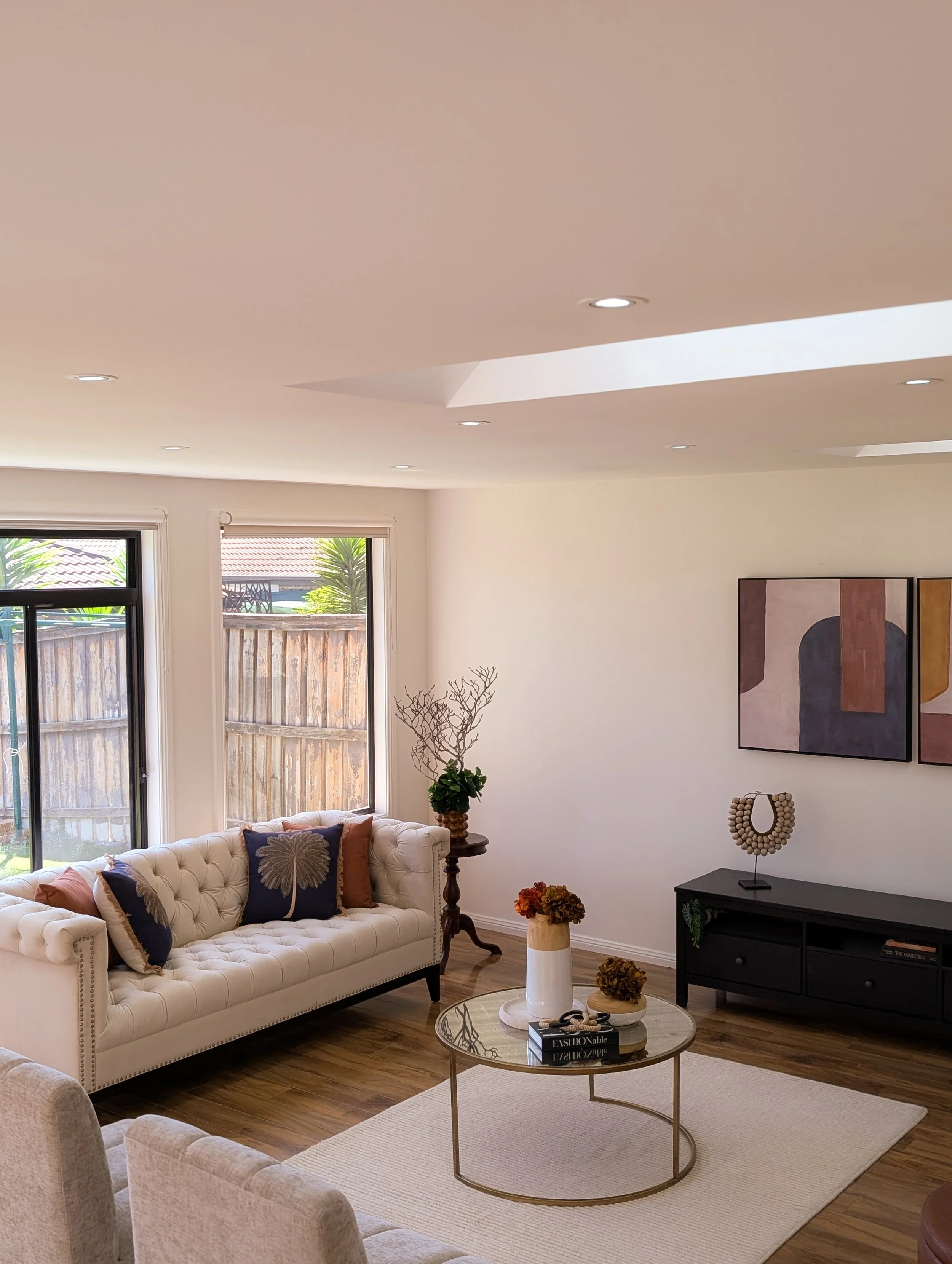 Living room with a beige tufted sofa with decorative pillows, a round glass coffee table with flowers and books, a black TV stand, and abstract artwork on a white wall. Two large windows with white blinds and a wooden fence outside.