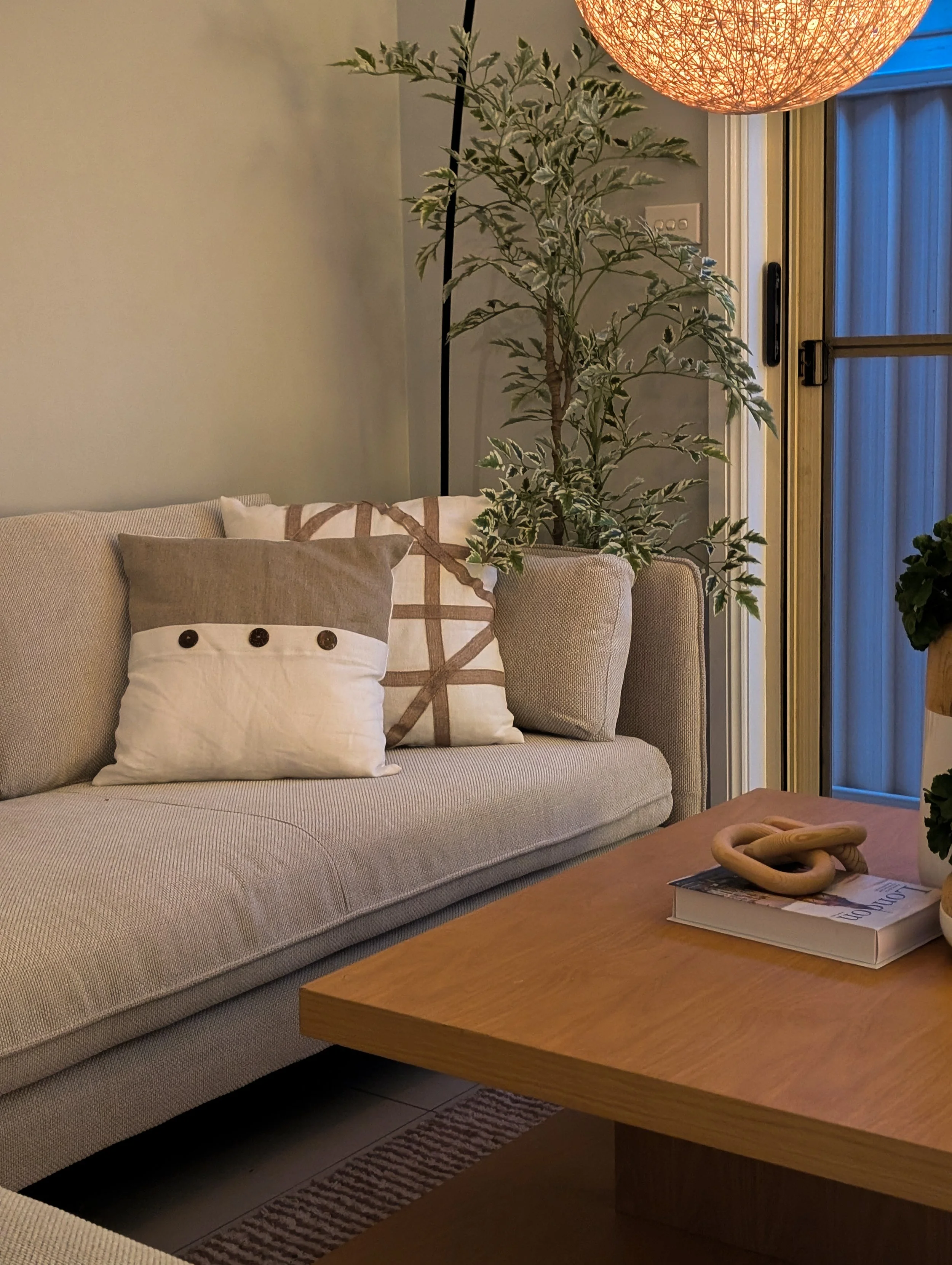 A cozy living room corner with a beige sofa decorated with patterned and solid pillows, a tall potted plant, a wooden coffee table with a book, a decorative knot, and a hanging woven light fixture.