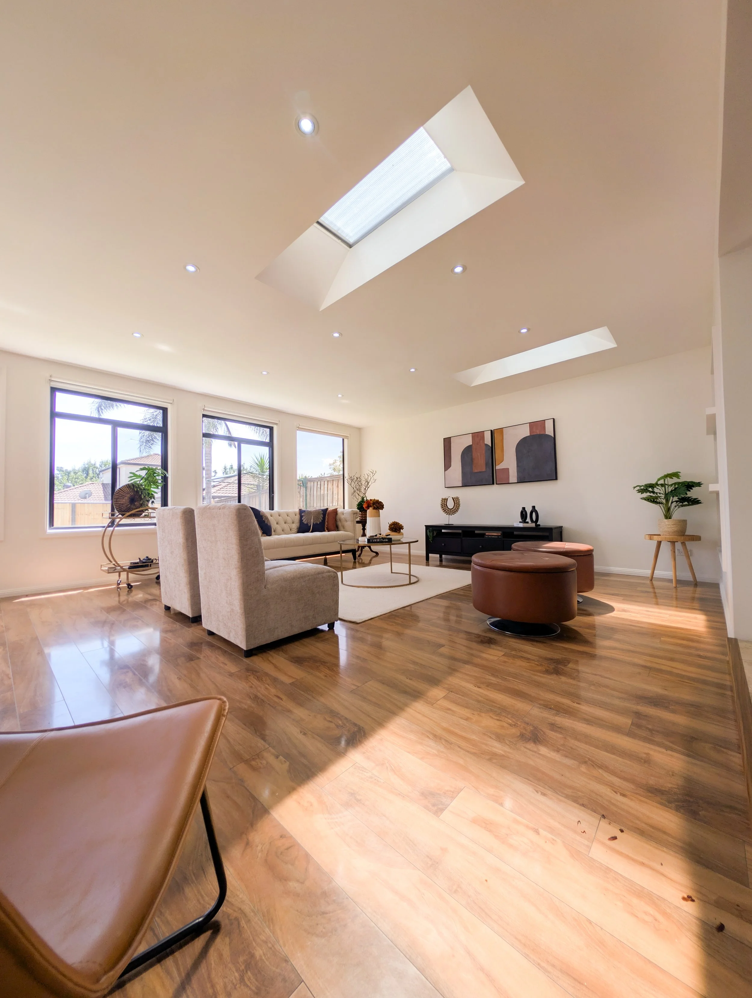 Bright living room with wood flooring, large windows, beige sofa, two beige armchairs, a black TV stand, abstract wall art, and skylights in the ceiling.