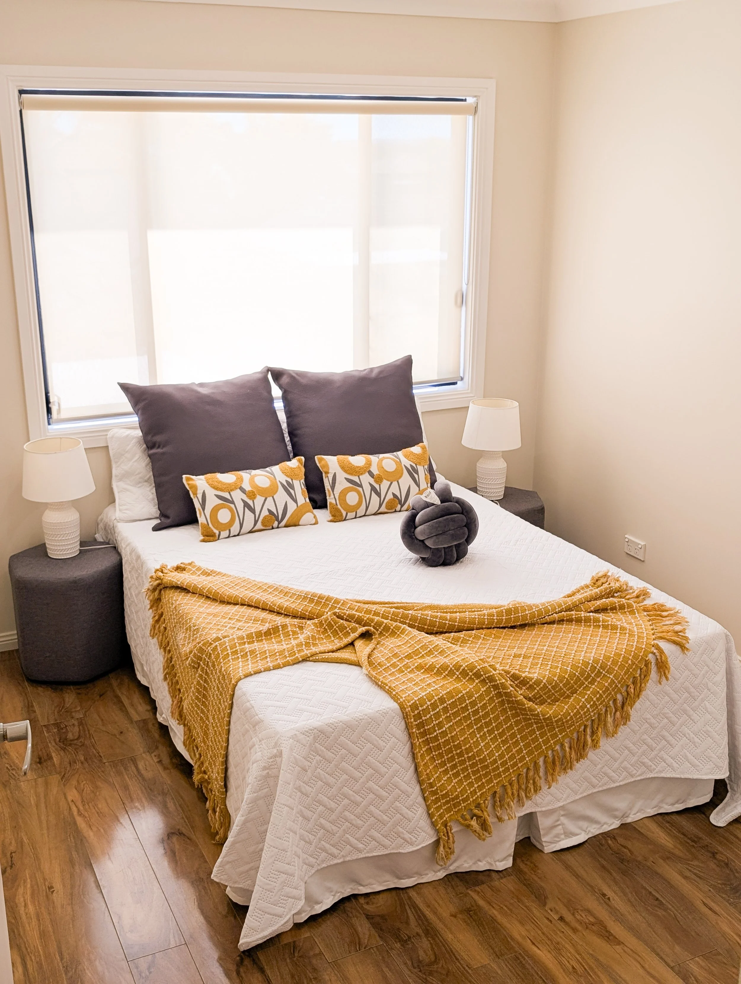 A neatly made bedroom with a large window, two end tables with lamps, decorative pillows, and a yellow throw blanket on the bed.