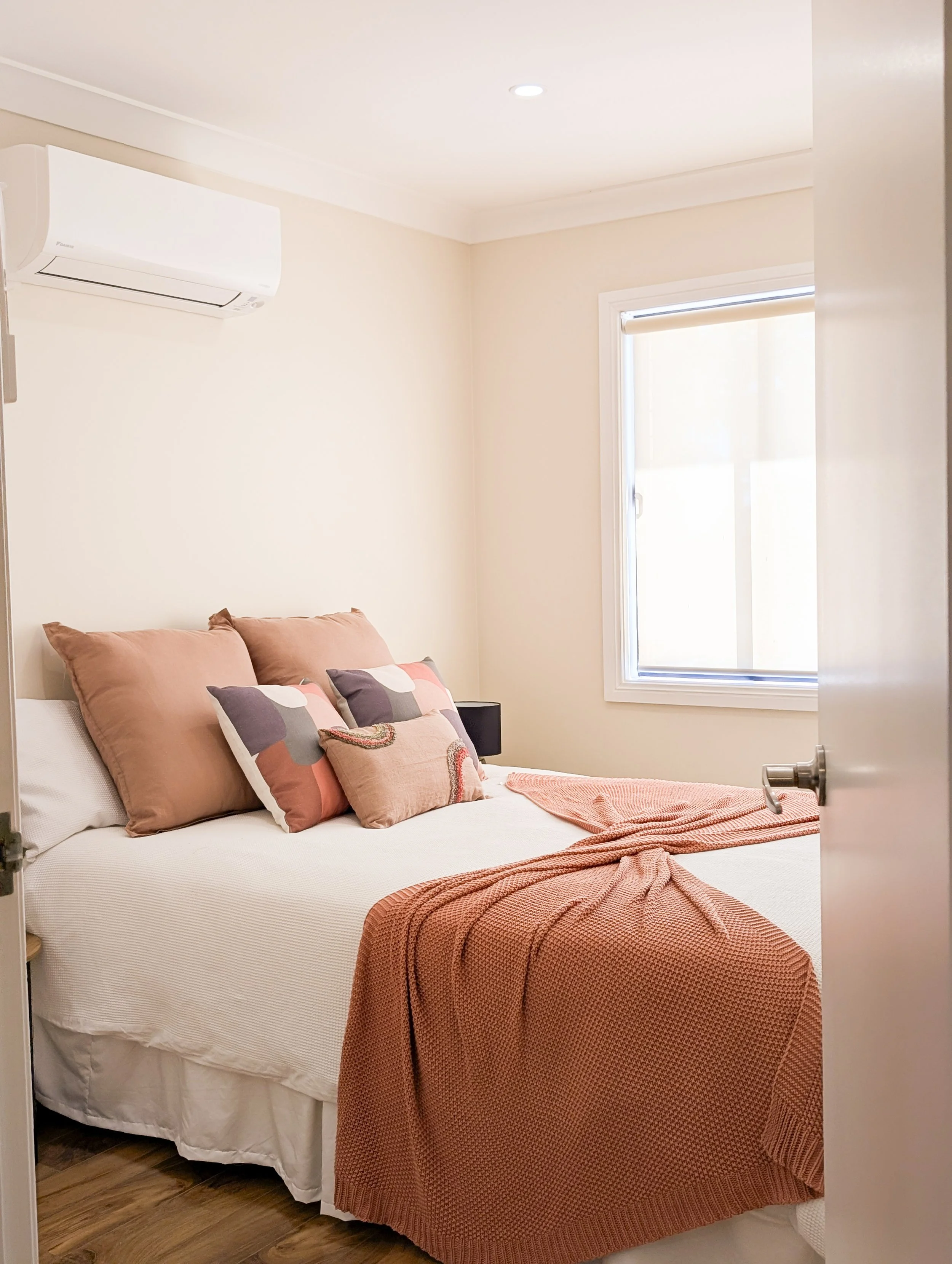 A cozy bedroom with a bed covered in a peach blanket, with multiple pillows, near a window with closed blinds and an air conditioning unit on the wall.