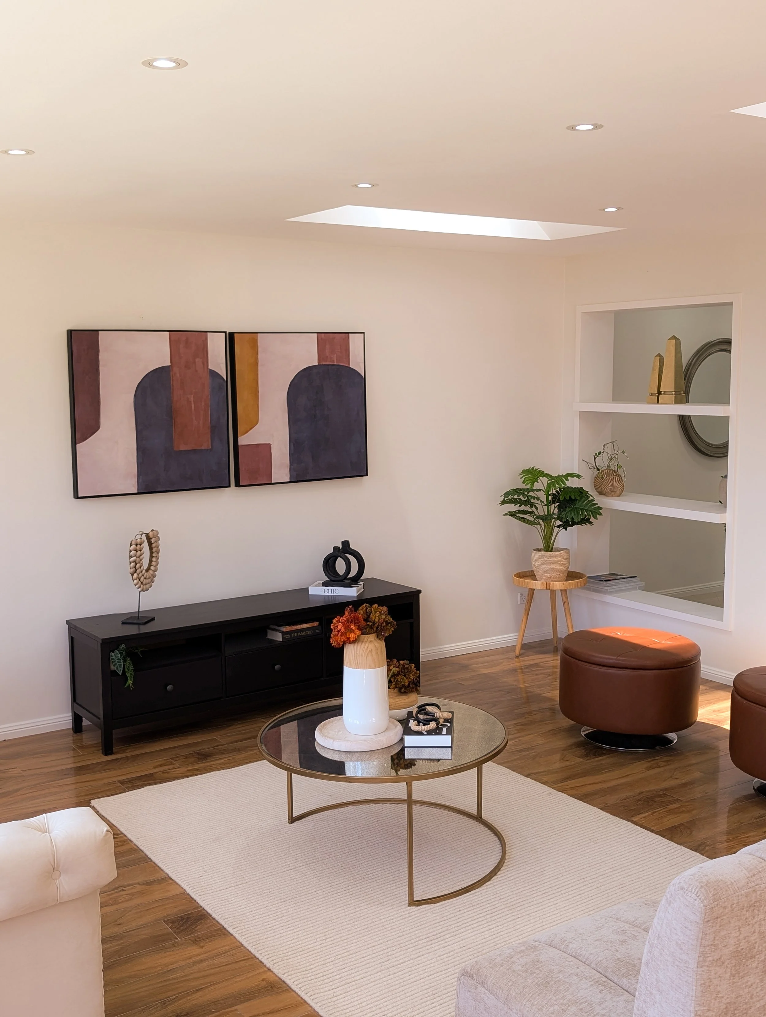 A modern living room with abstract art on the wall, a black TV stand, a glass-top coffee table with decorative items, a beige rug, a beige sofa, brown ottomans, and various plants and decorations.