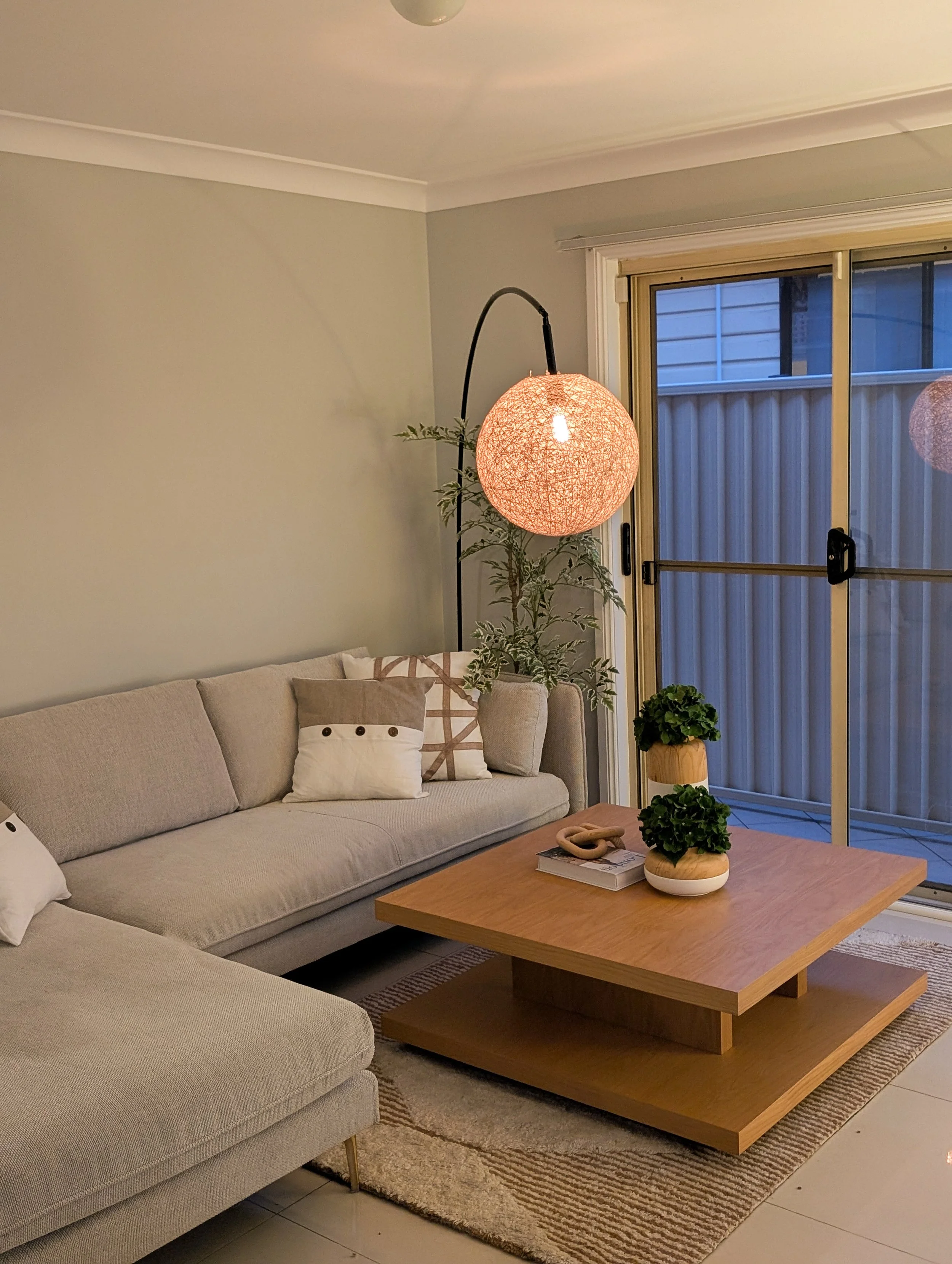 Living room with a beige sectional sofa, decorative pillows, a wooden coffee table with potted plants, a floor lamp with a round pink shade, and a sliding glass door leading outside.