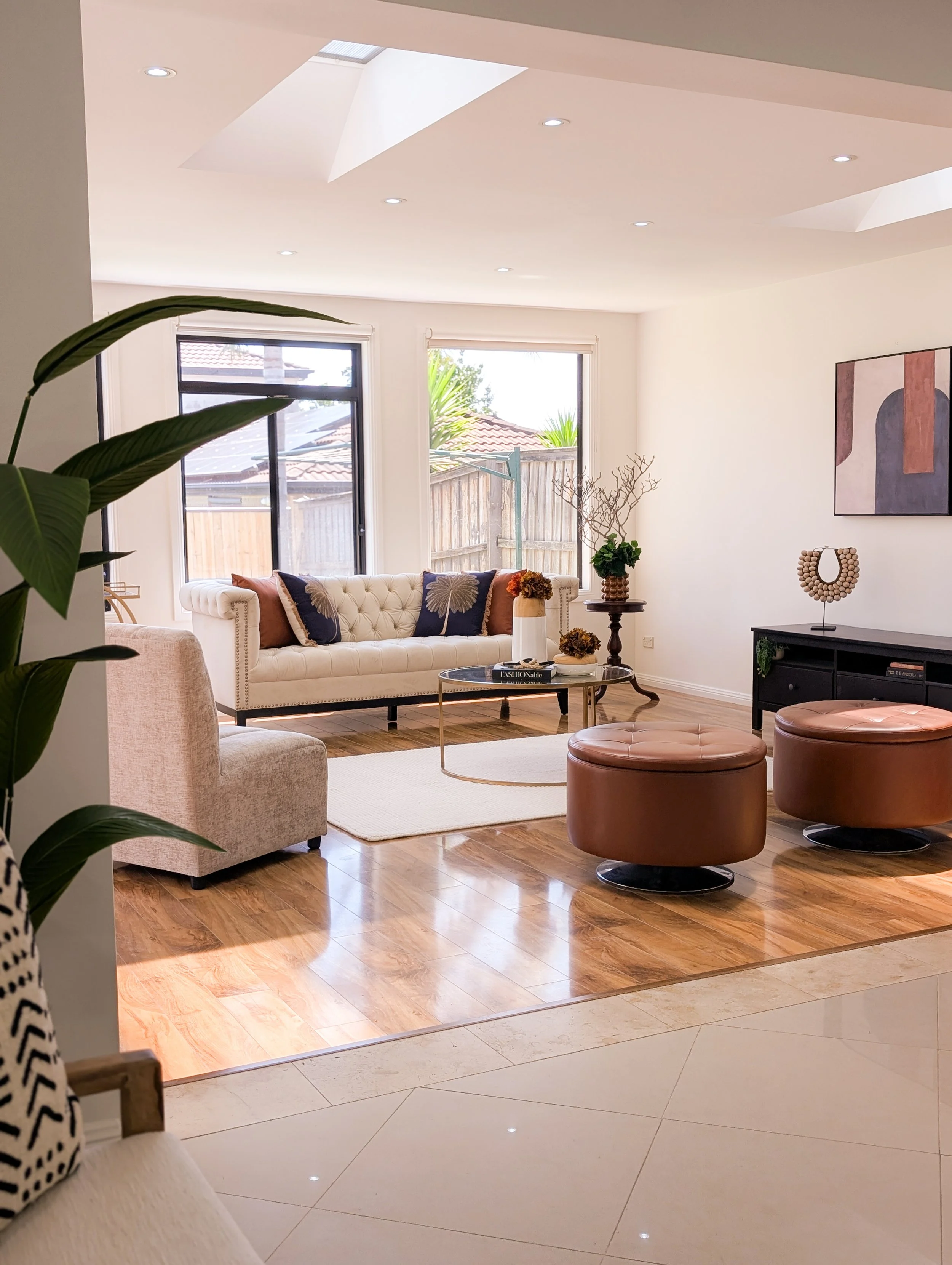 Bright living room with white tufted sofa, beige armchair, brown ottomans, glass coffee table, black sideboard, large windows, and decorative plants.