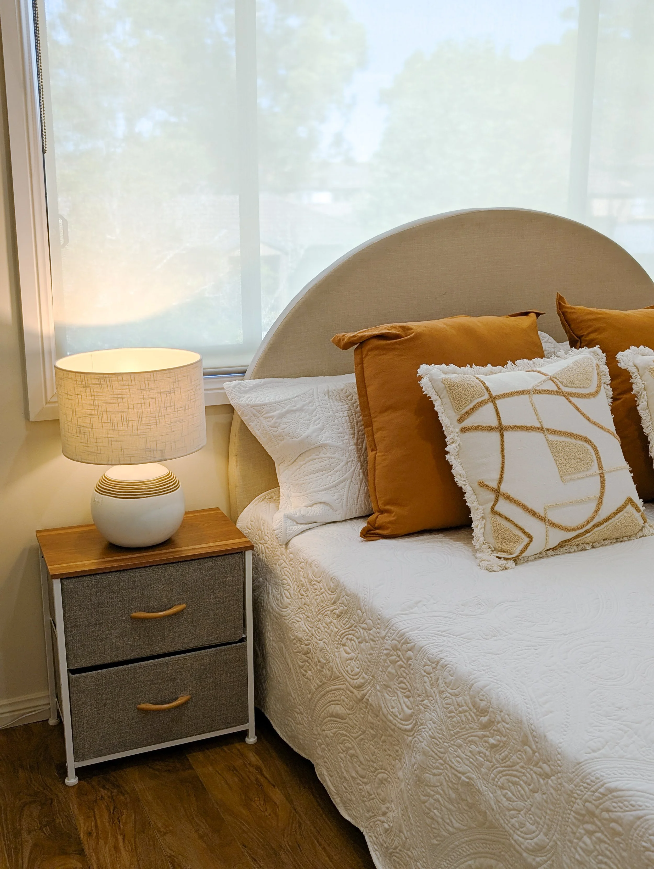 A bedroom with a beige upholstered headboard, white textured bedspread, decorative pillows in shades of brown and white with fringe, a wooden nightstand with two fabric drawers, a round ceramic table lamp, a window with frosted glass and sheer curtai