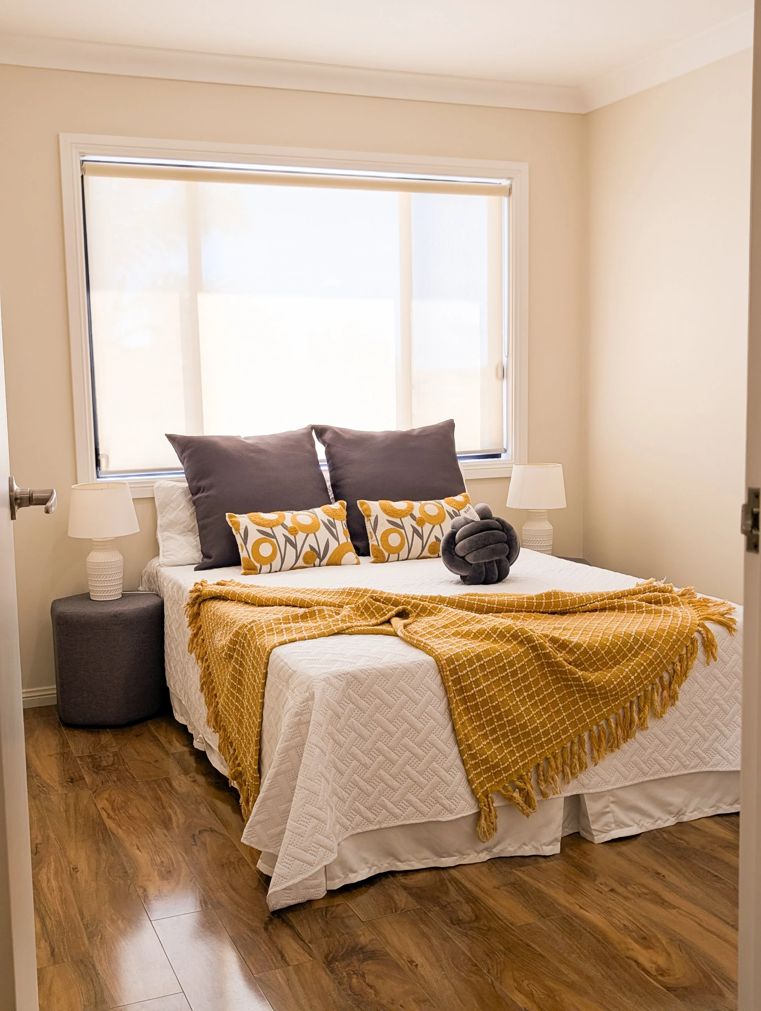 A bedroom with a bed, two pillows, two decorative pillows, and an orange blanket. There are two nightstands with lamps and a window with closed blinds.