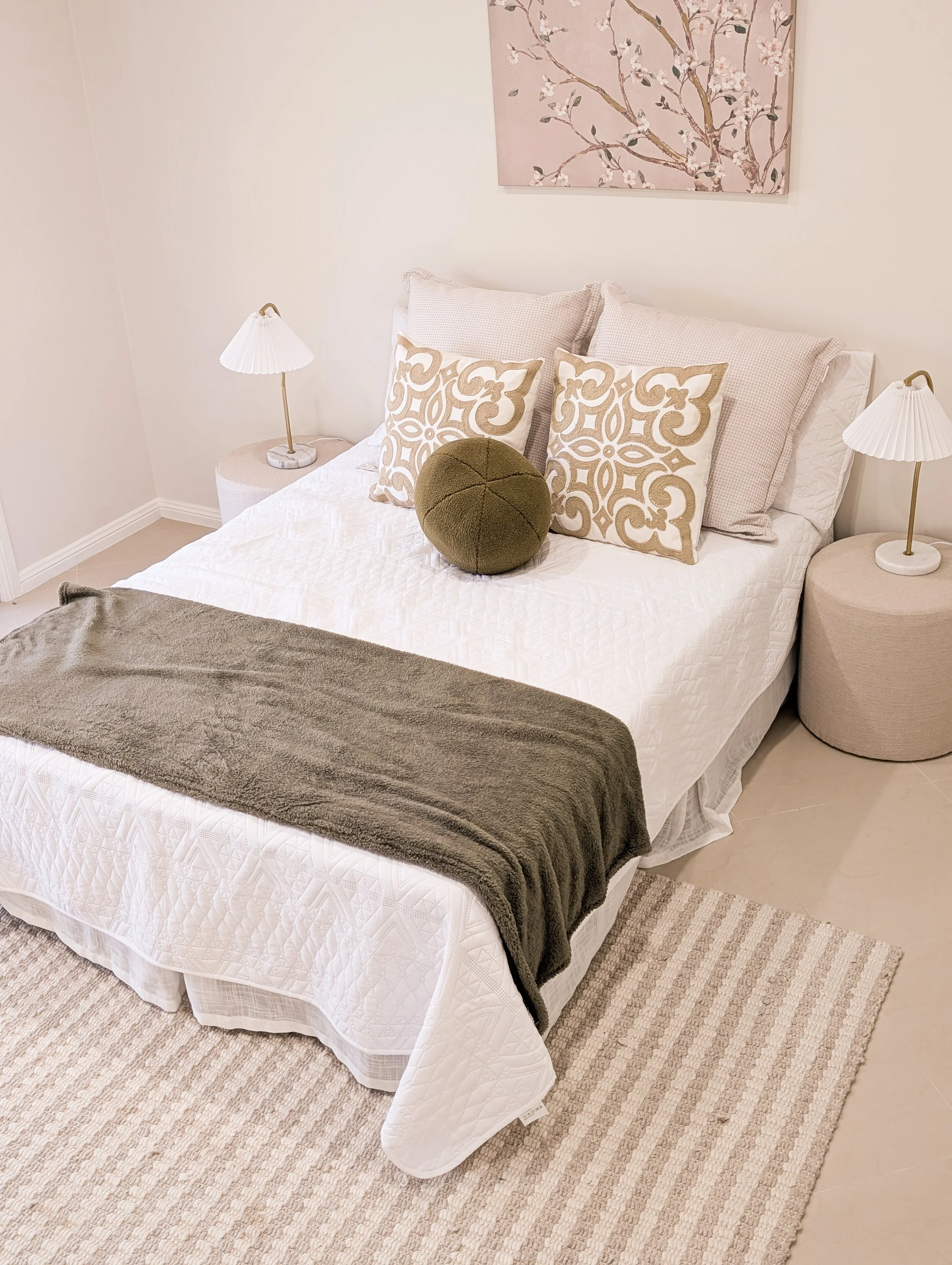 A bedroom with a neatly made bed, decorative pillows, a green round pillow, beige bedside tables with white lamps, a pink painting of cherry blossoms, and a beige rug.