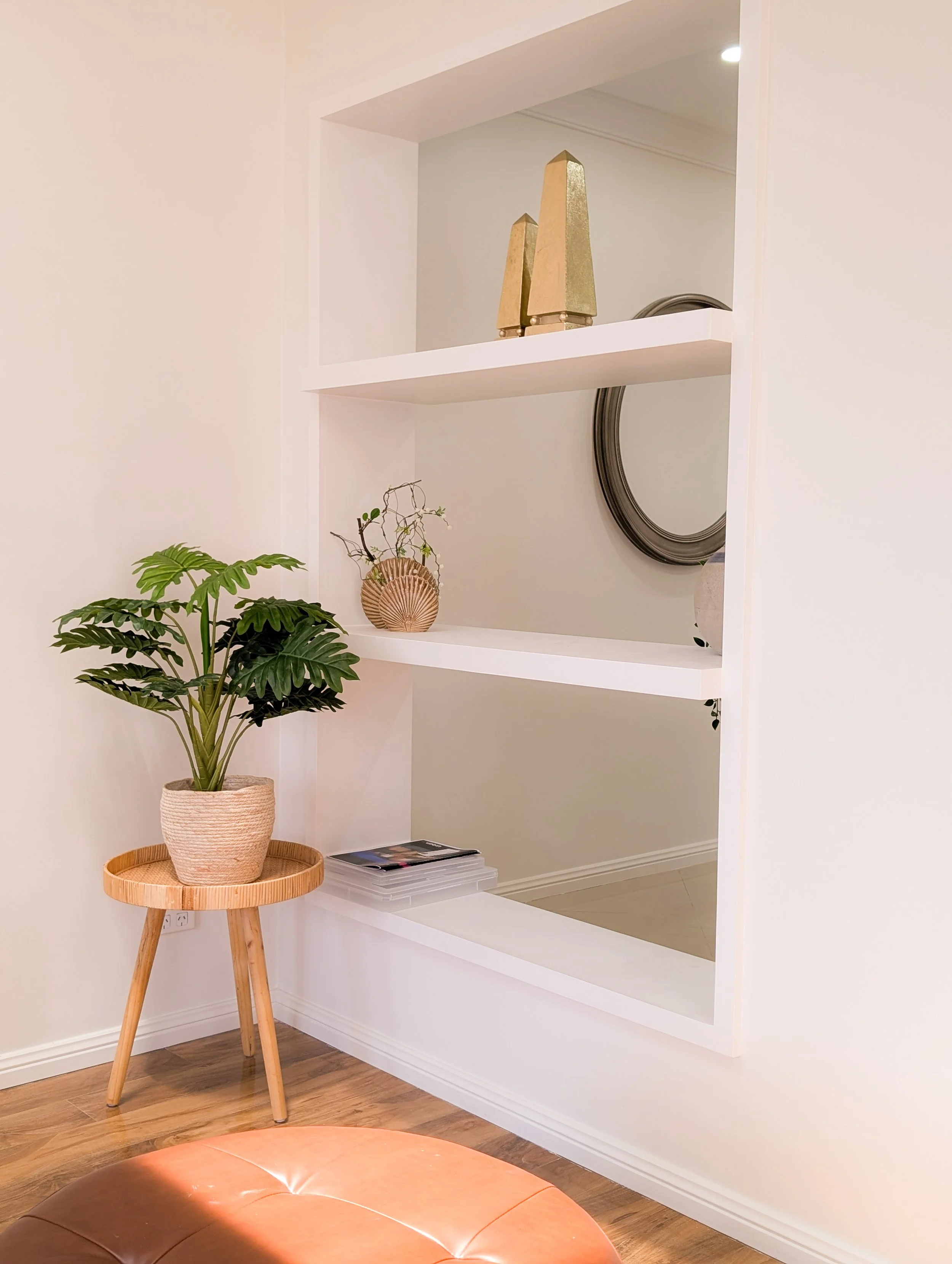 Interior corner with white built-in shelves, a potted plant on a wooden stand, and decorative items on the shelves including gold obelisks, a mirror, and a small basket with branches.