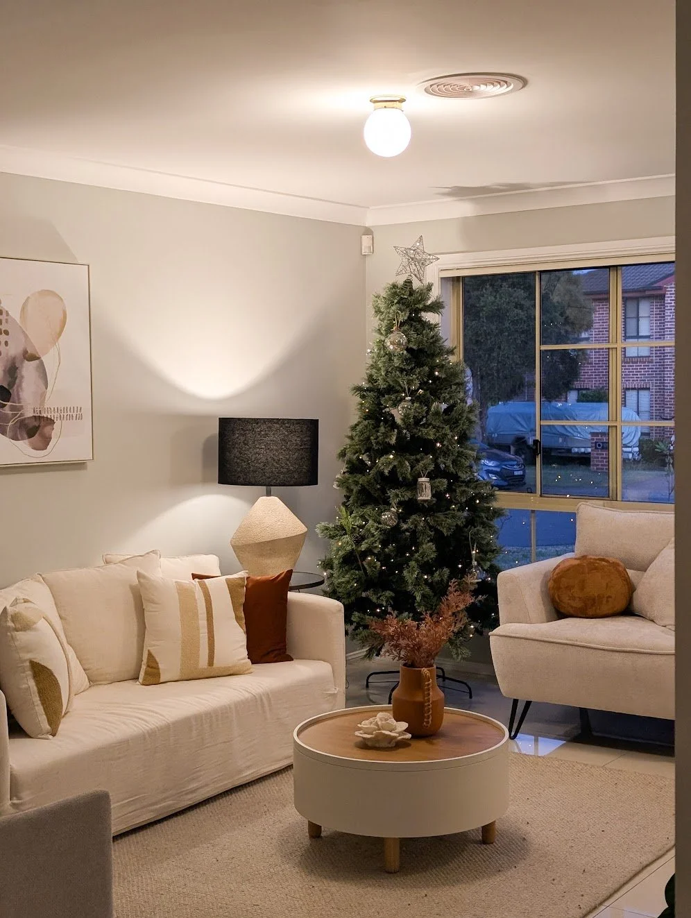 Living room decorated for Christmas with a decorated Christmas tree, a beige sofa with pillows, a side table with a black lamp, a round coffee table with a vase and decorative objects, and a large window showing a street scene outside.