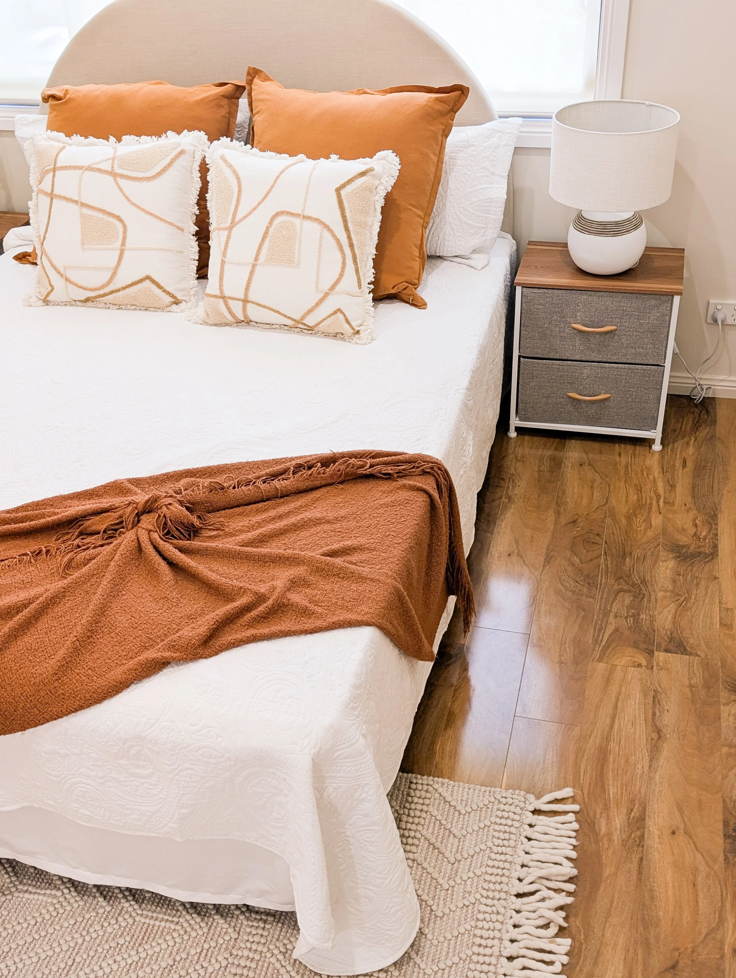 A neatly made bed with white quilted bedding, two rust-colored pillows, and two decorative white pillows with abstract orange lines. A rust-colored blanket is draped over the foot of the bed. To the right, a wooden nightstand with two drawers and a c