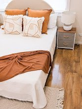 A neatly made bed with white bedding, orange and beige throw pillows, and a rust-colored blanket at the foot. There is a gray nightstand with a small decorative object and a white lamp on it. The room has wood flooring and a beige rug.