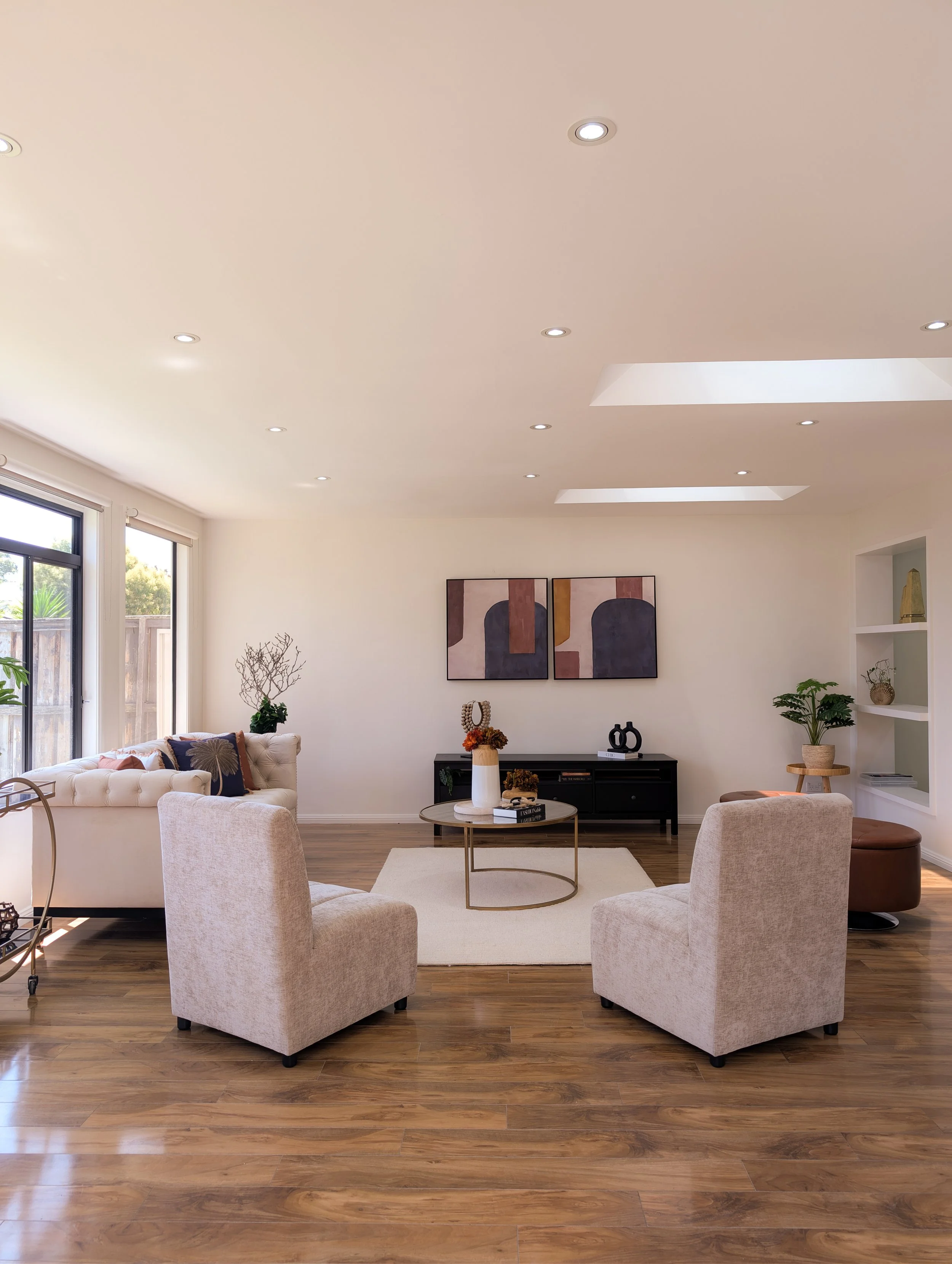 Modern living room with beige chairs, white sofa, wooden floor, coffee table, wall art, and large windows.