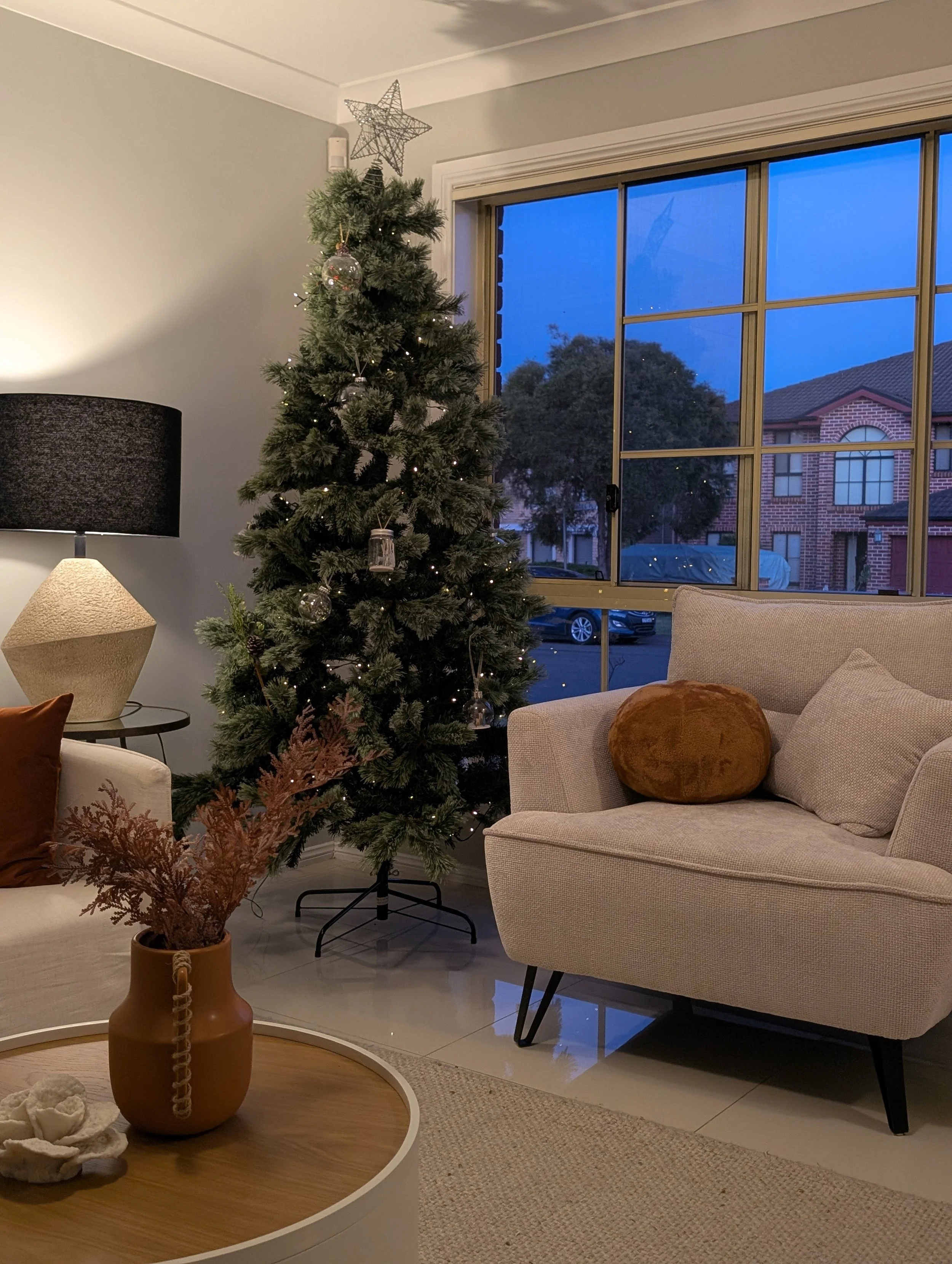 Interior of a living room decorated for Christmas, featuring a Christmas tree with ornaments and lights, a beige sofa with a round brown cushion, a round wooden coffee table with a vase and decorative items, and a window showing a neighborhood scene 
