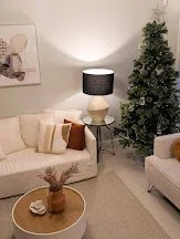 Living room with beige sofa, round gold coffee table, potted plant, and Christmas tree. Wall art and table lamp included.
