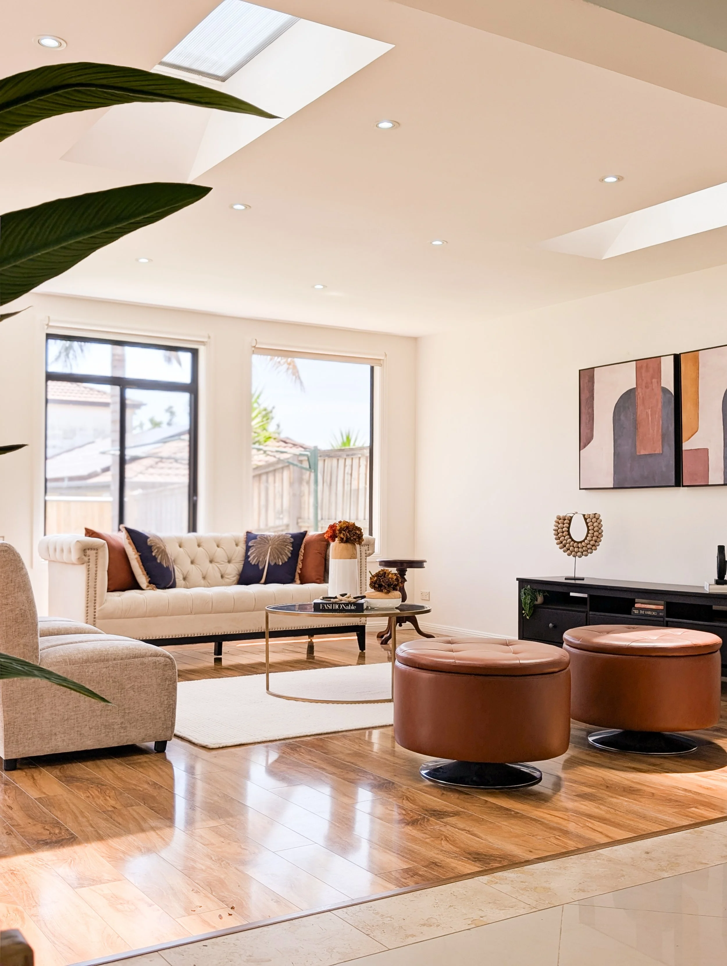 Modern living room with beige tufted sofa, armchair, two brown leather ottomans, coffee table, wall art, and large windows with wooden fencing outside.