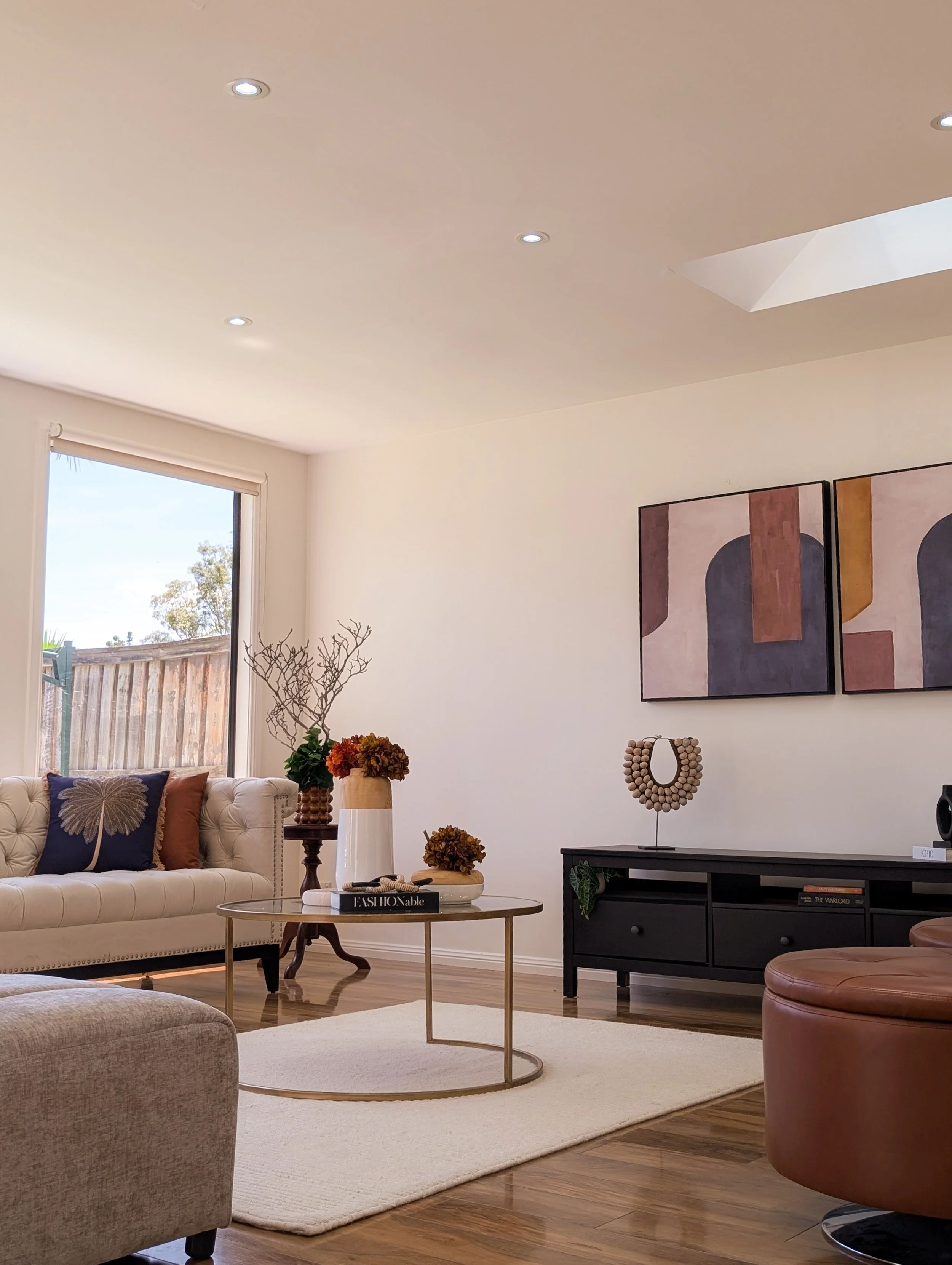 Modern living room with beige tufted sofa, armchair, black TV console, abstract wall art, round coffee table, decorative vases, and minimal decor.
