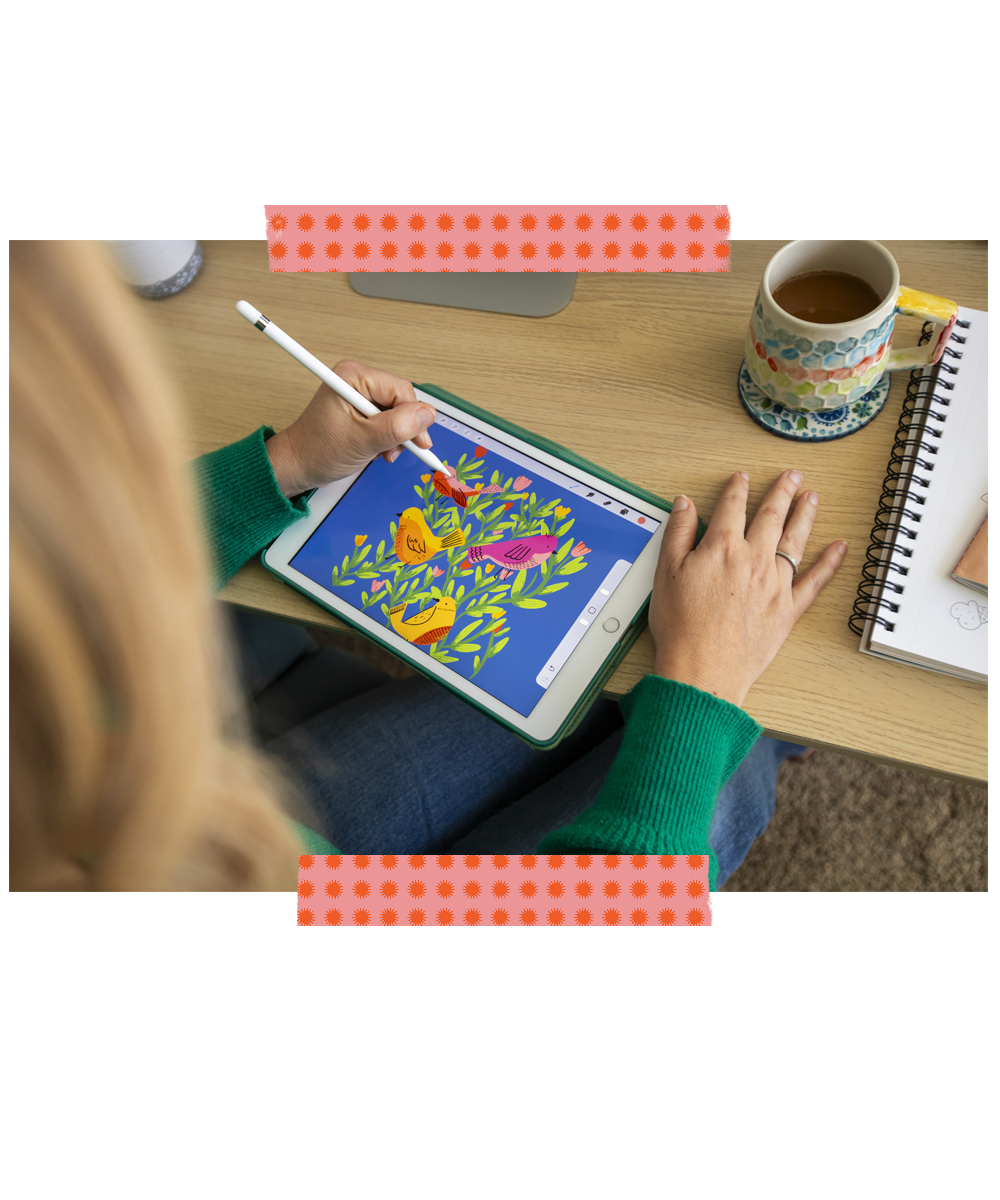 An illustrator drawing colorful birds and flowers on a digital tablet with a stylus at a wooden desk, with a cup of tea, a notebook, and a drink in the background.