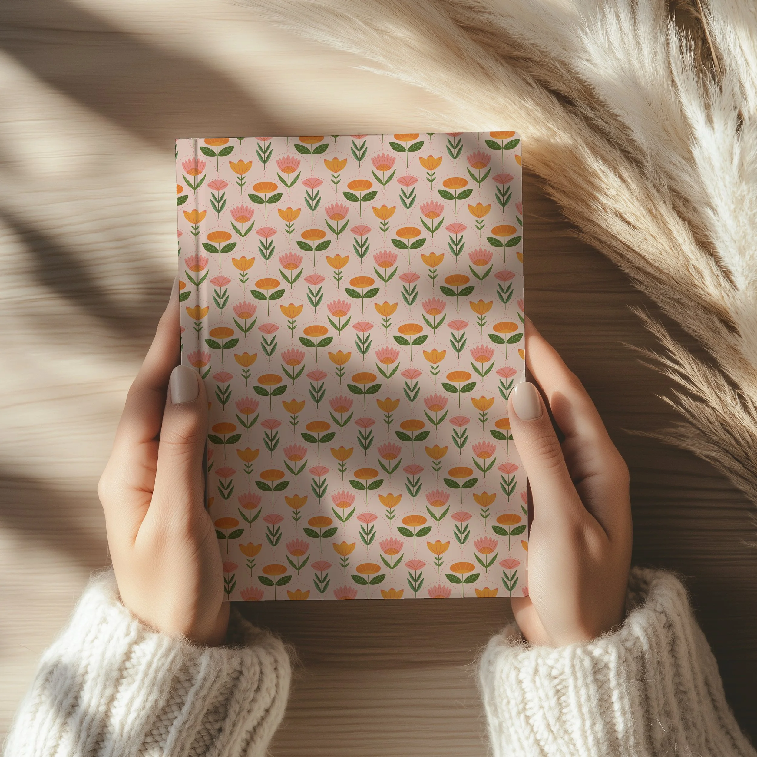 A person holding a patterned journal with pink, orange, and yellow flowers and green leaves. The person is wearing a white knit sweater, and there are soft shadows and dried plants on a light wooden surface.