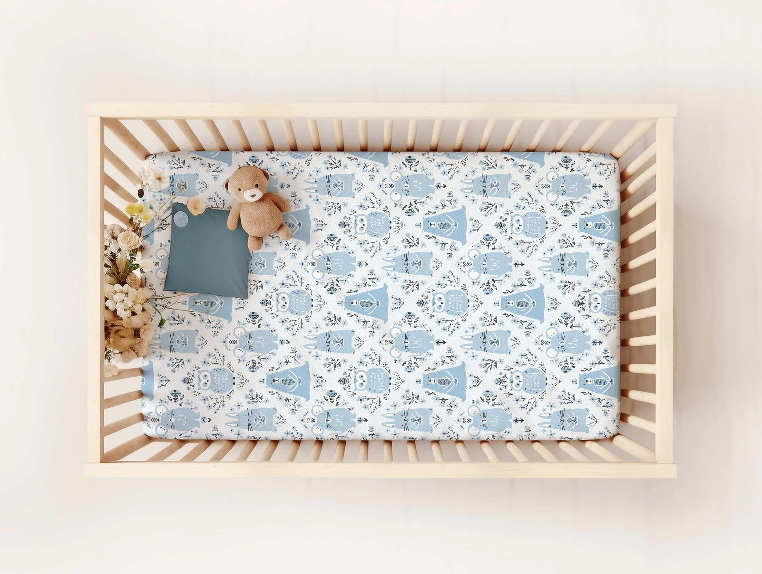 Top-down view of a wooden baby crib with a blue animal-themed mattress, a small teddy bear, a blue pillow, and a bunch of flowers on the corner.