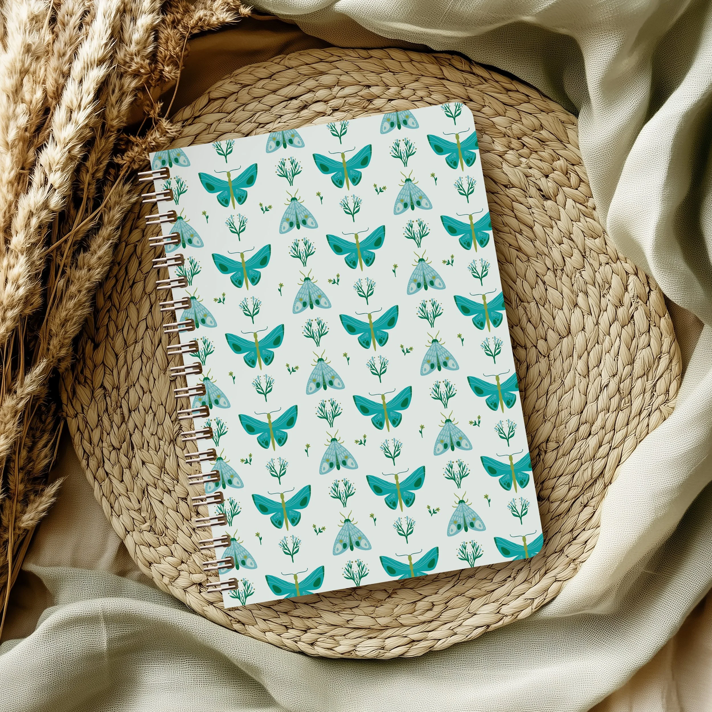 Notebook with butterfly and plant pattern on a woven placemat, surrounded by dried wheat stalks and fabric.